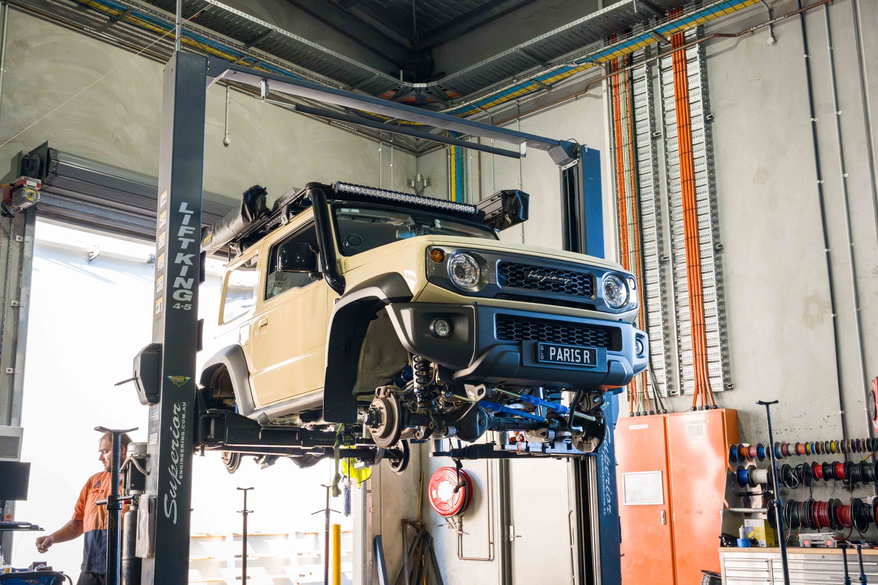 Beige off-road SUV elevated on a two‑post lift inside a workshop, technician in orange nearby inspecting it.