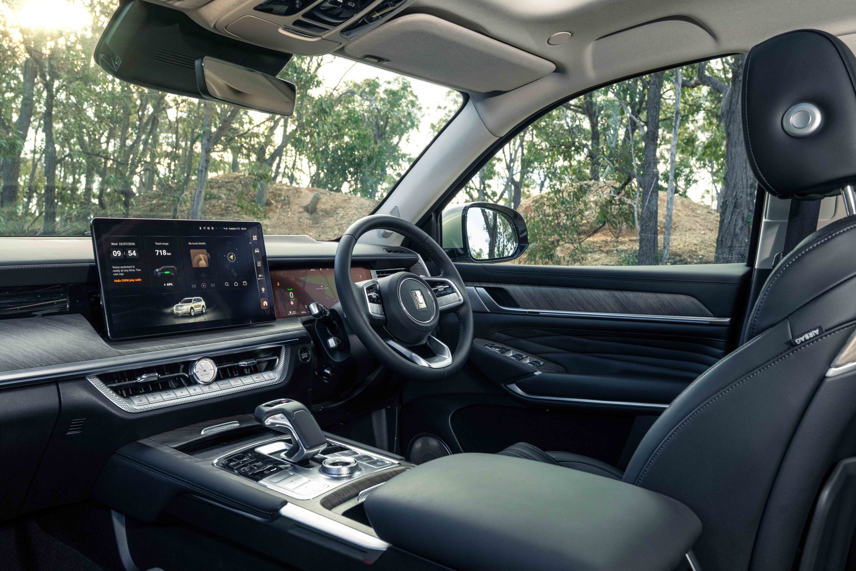 Luxurious car interior showing a wide central touchscreen display, steering wheel, and polished center console with chrome accents.Outside view of trees through the passenger window.