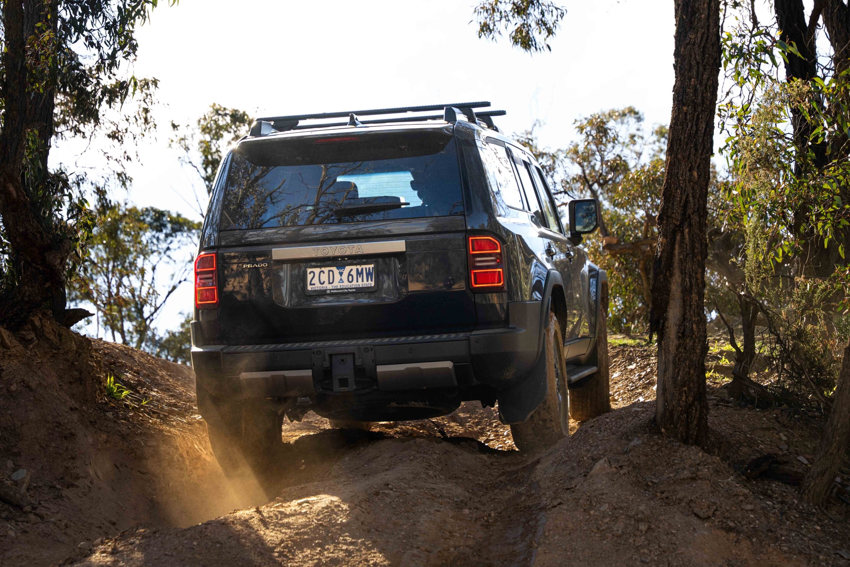 Black Toyota Prado driving on a rocky dirt trail, dust rising as it descends between trees
