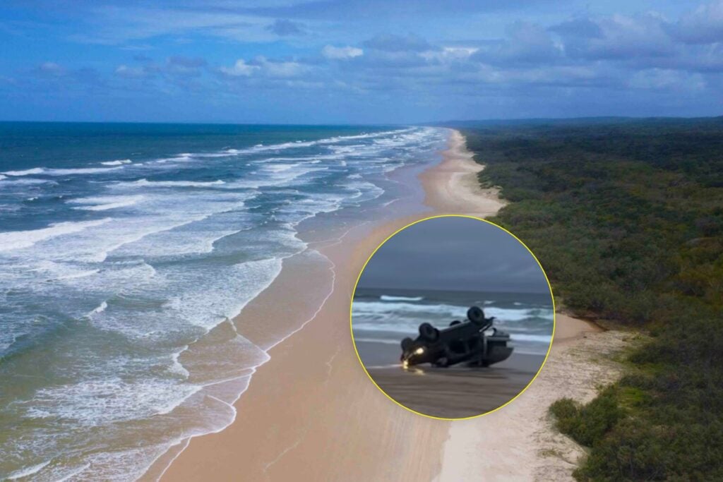 Aerial view of a long sandy beach with blue ocean and a forested coastline; an inset circle shows an overturned vehicle on a wet road.