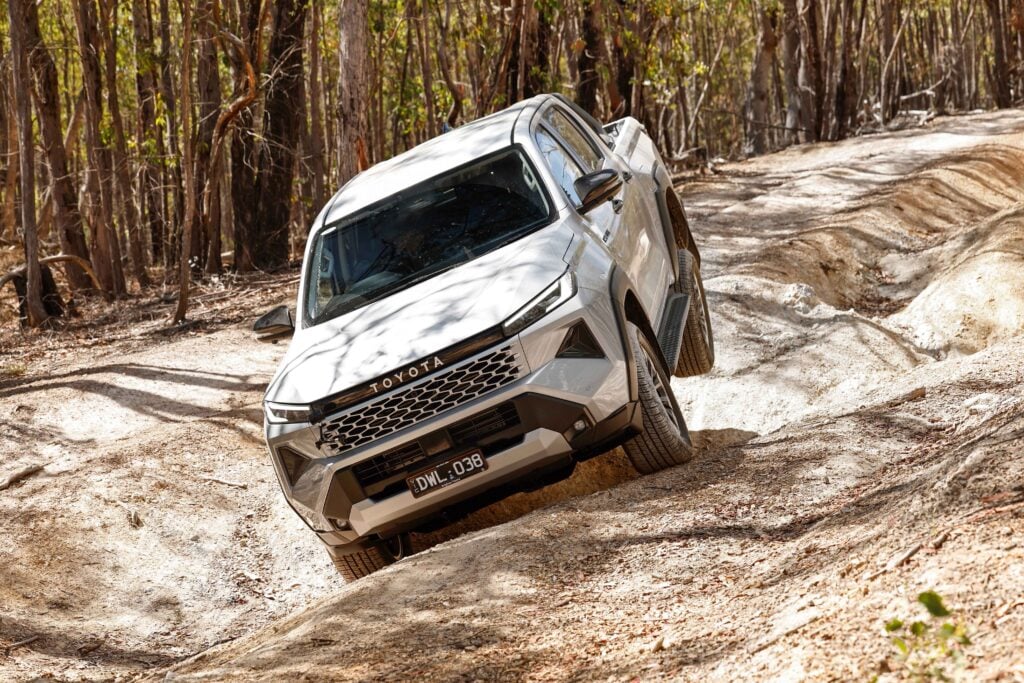 White Toyota SUV on a rough dirt trail in a forest, tilted as it climbs an uneven incline.