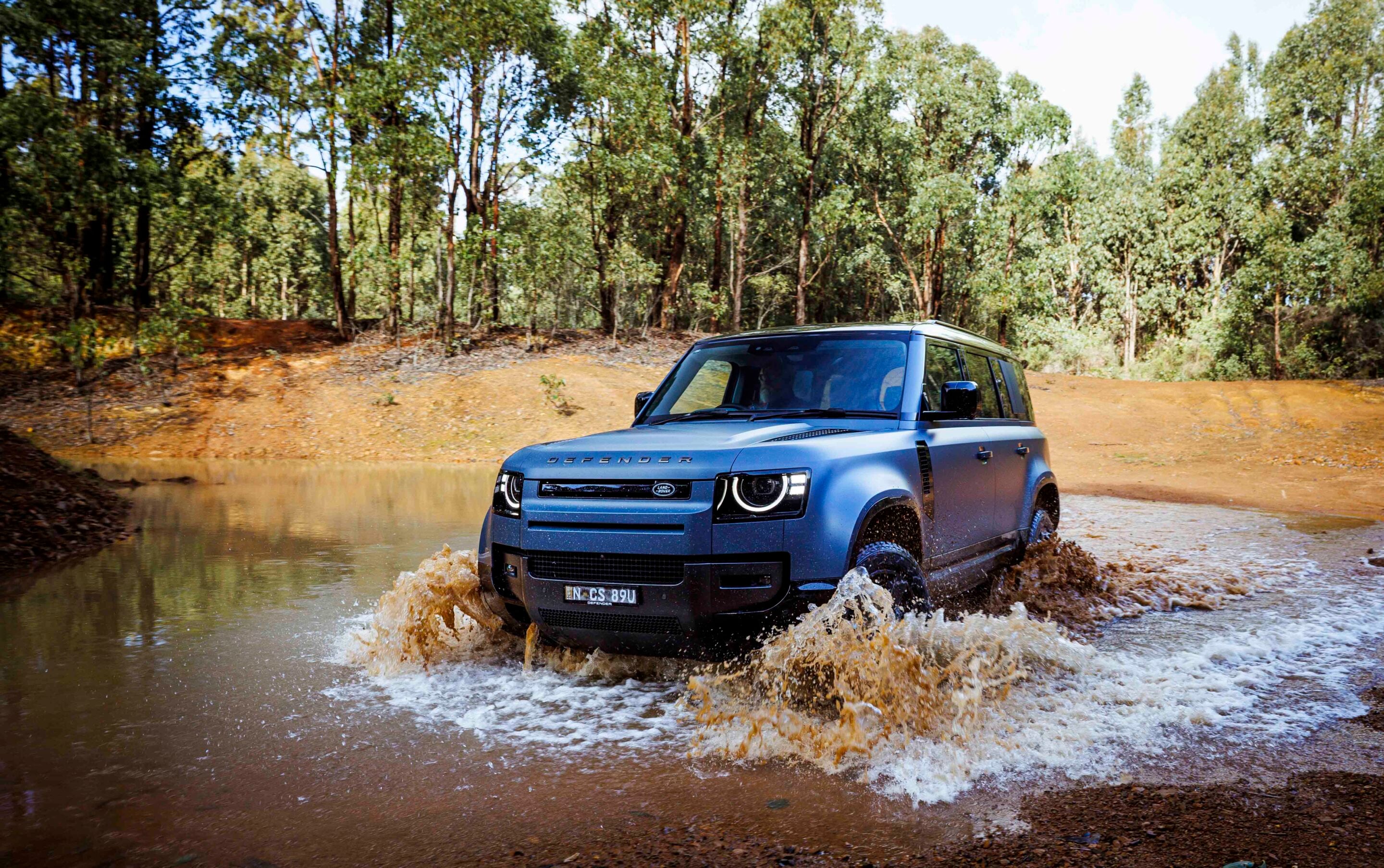 Blue Land Rover Defender driving through muddy water in a forest, splashing as it passes.