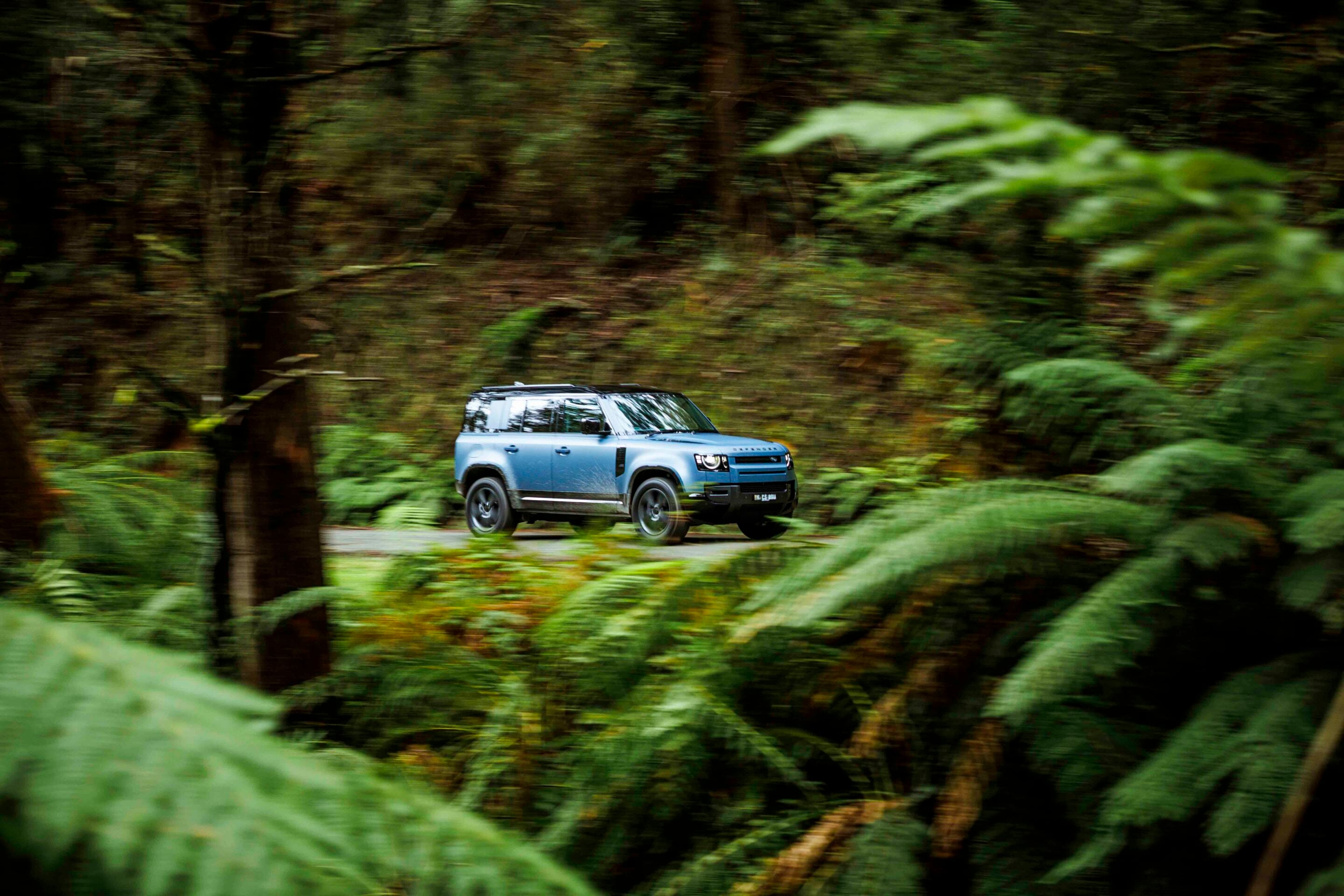 Blue Range Rover driving through a dense forest on a winding road, motion blurred to show speed.