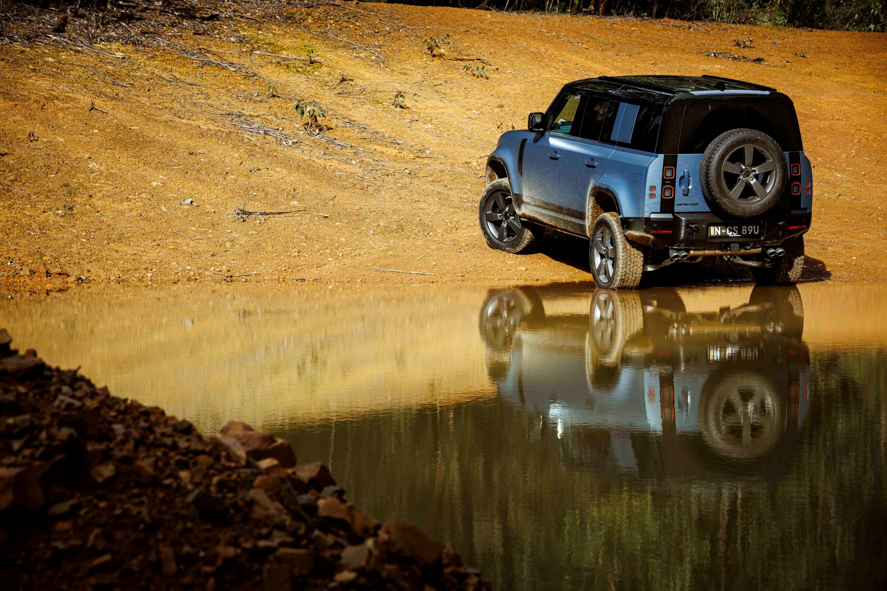 Blue off-road SUV (Land Rover Defender) parked on a muddy riverbank, with its reflection visible in the calm water nearby.