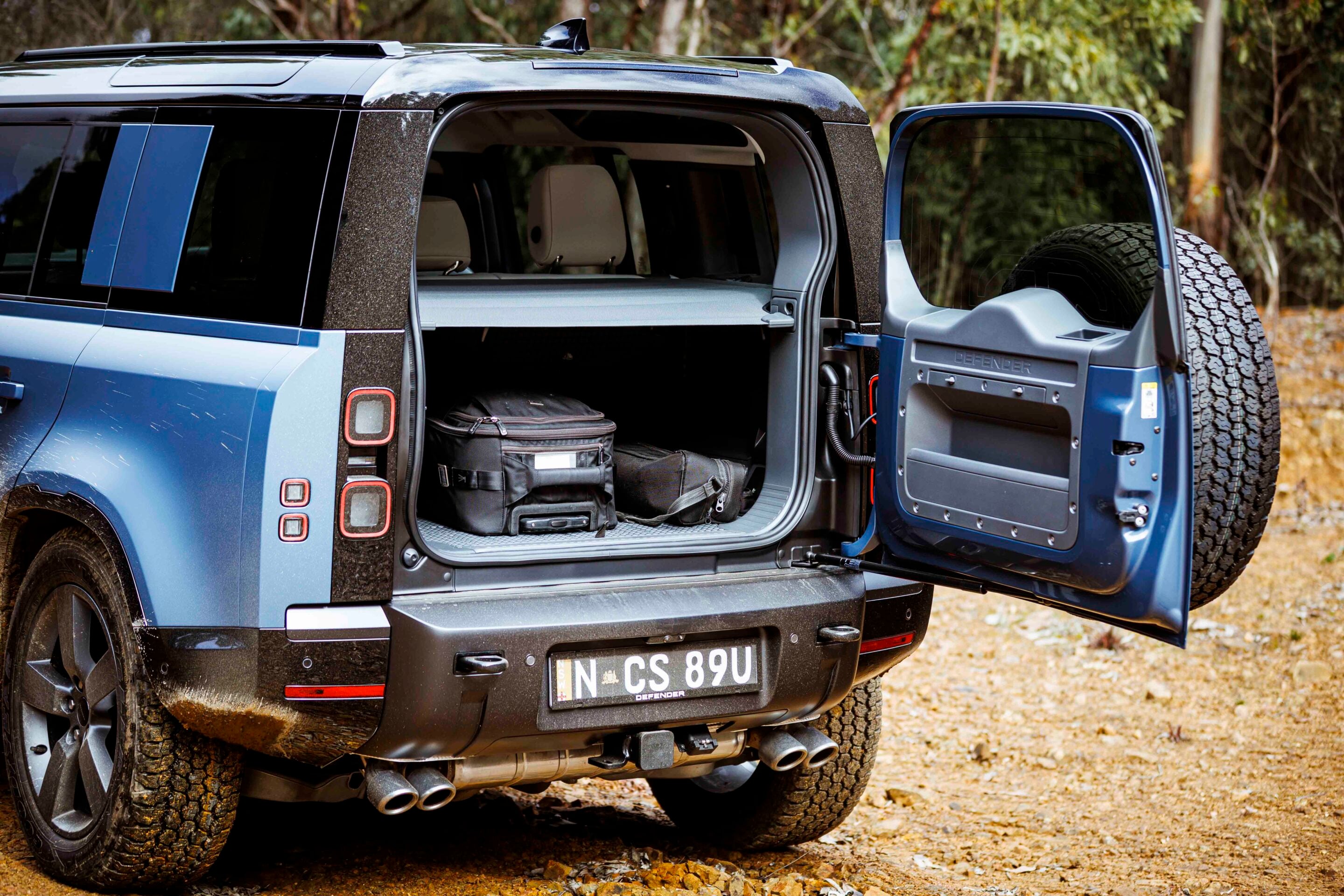 Rear of a blue Land Rover Defender with tailgate open, showing gear bags in the cargo area and a spare tire on the open door outside on a dirt path