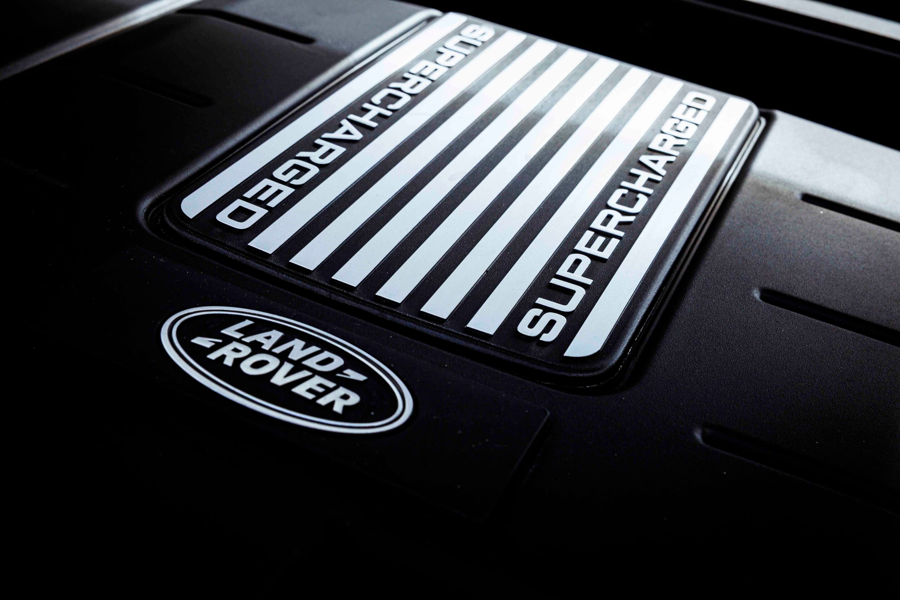 Close-up of a Land Rover badge on a black engine cover with chrome stripes and the word 'SUPERCHARGED' visible across the plate.