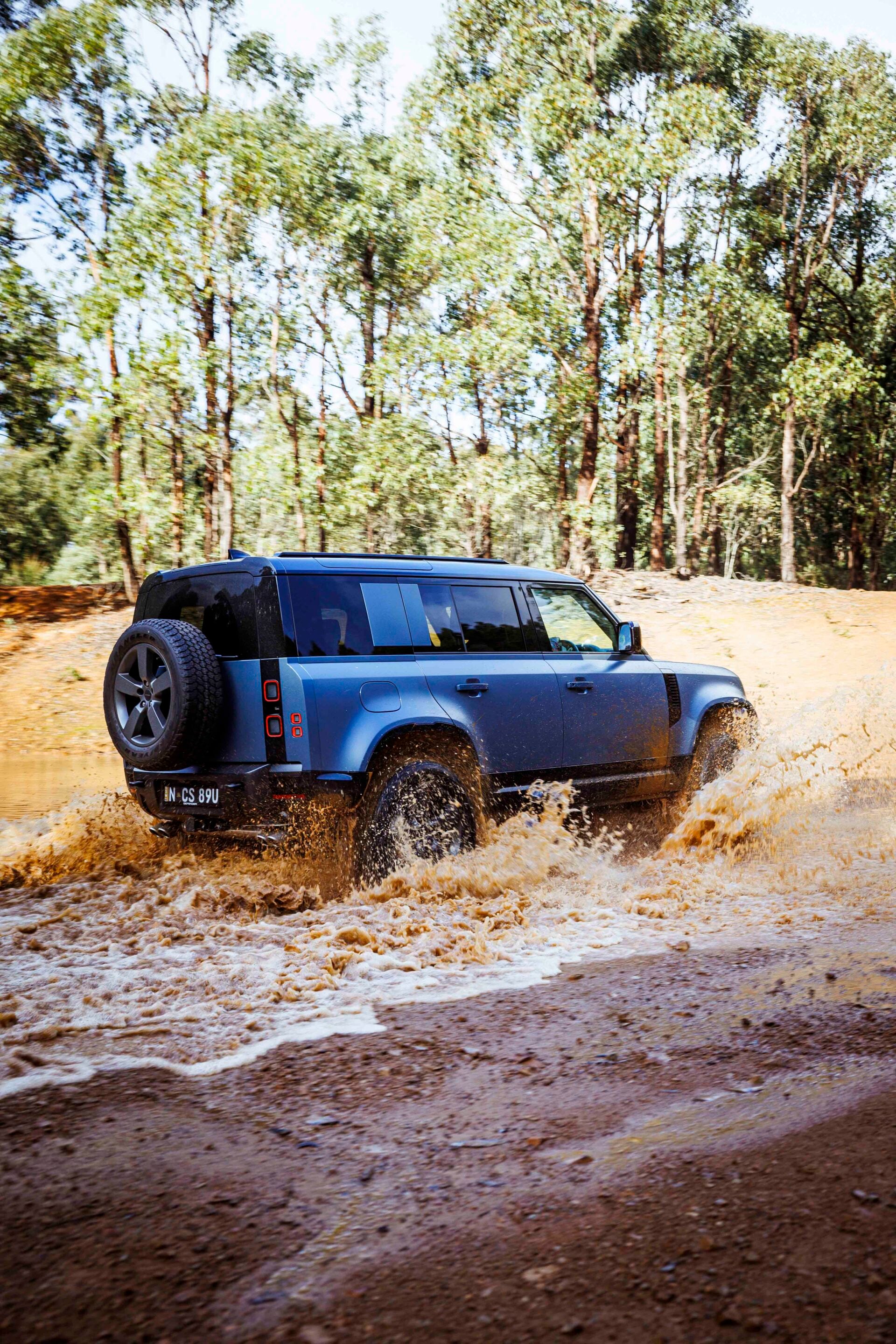 Blue SUV splashing through muddy water on a dirt road under trees in a forest setting.