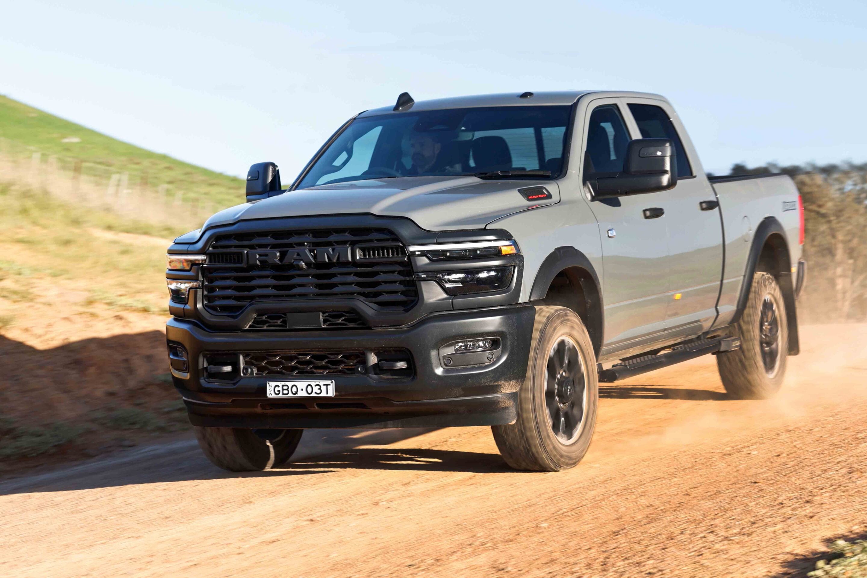 Front three-quarter view of a gray RAM pickup truck speeding along a dusty dirt road with hills in the background.