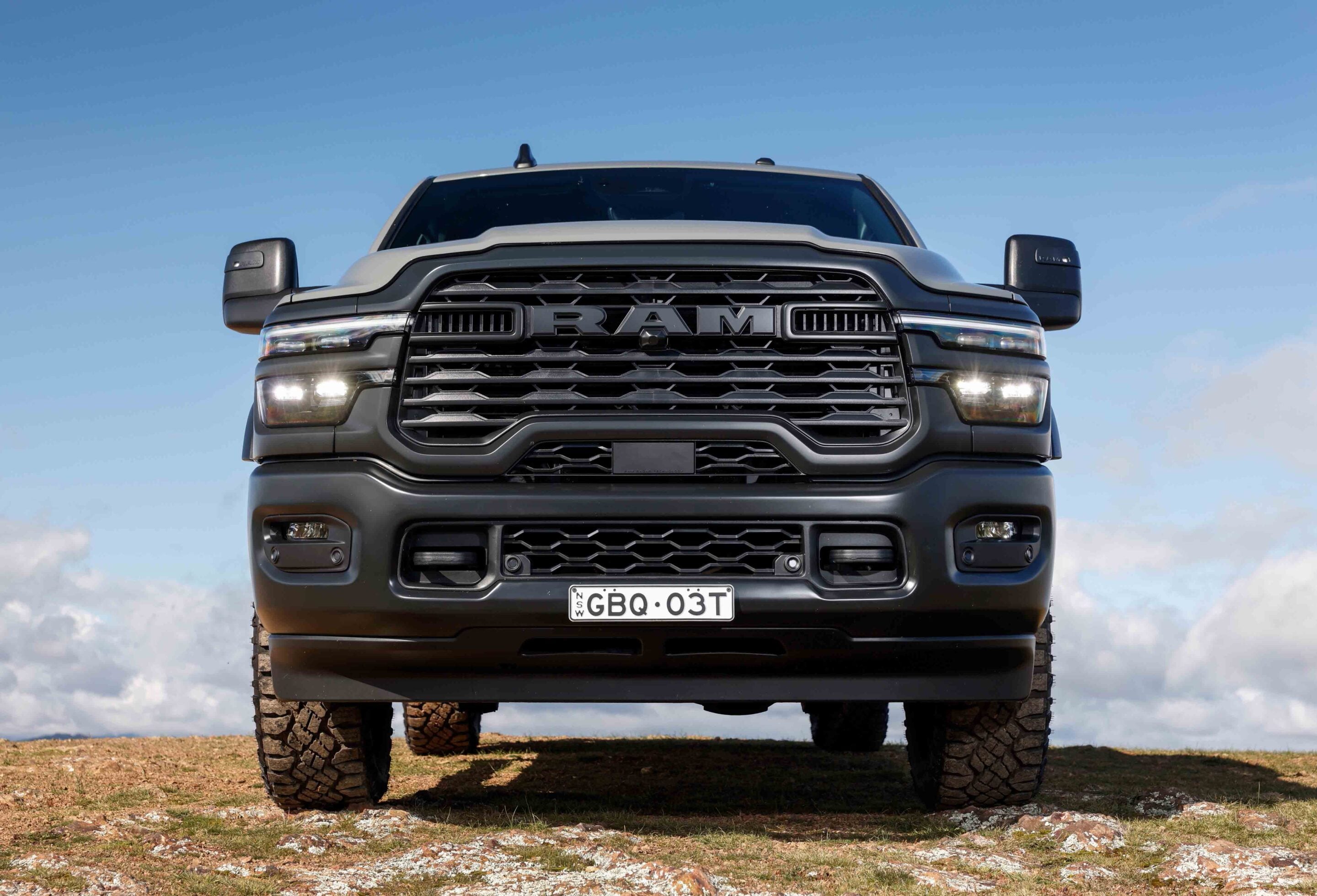 Front view of a gray RAM pickup truck on rocky ground, large grille with RAM logo and the plate GBQ-03T (New South Wales).