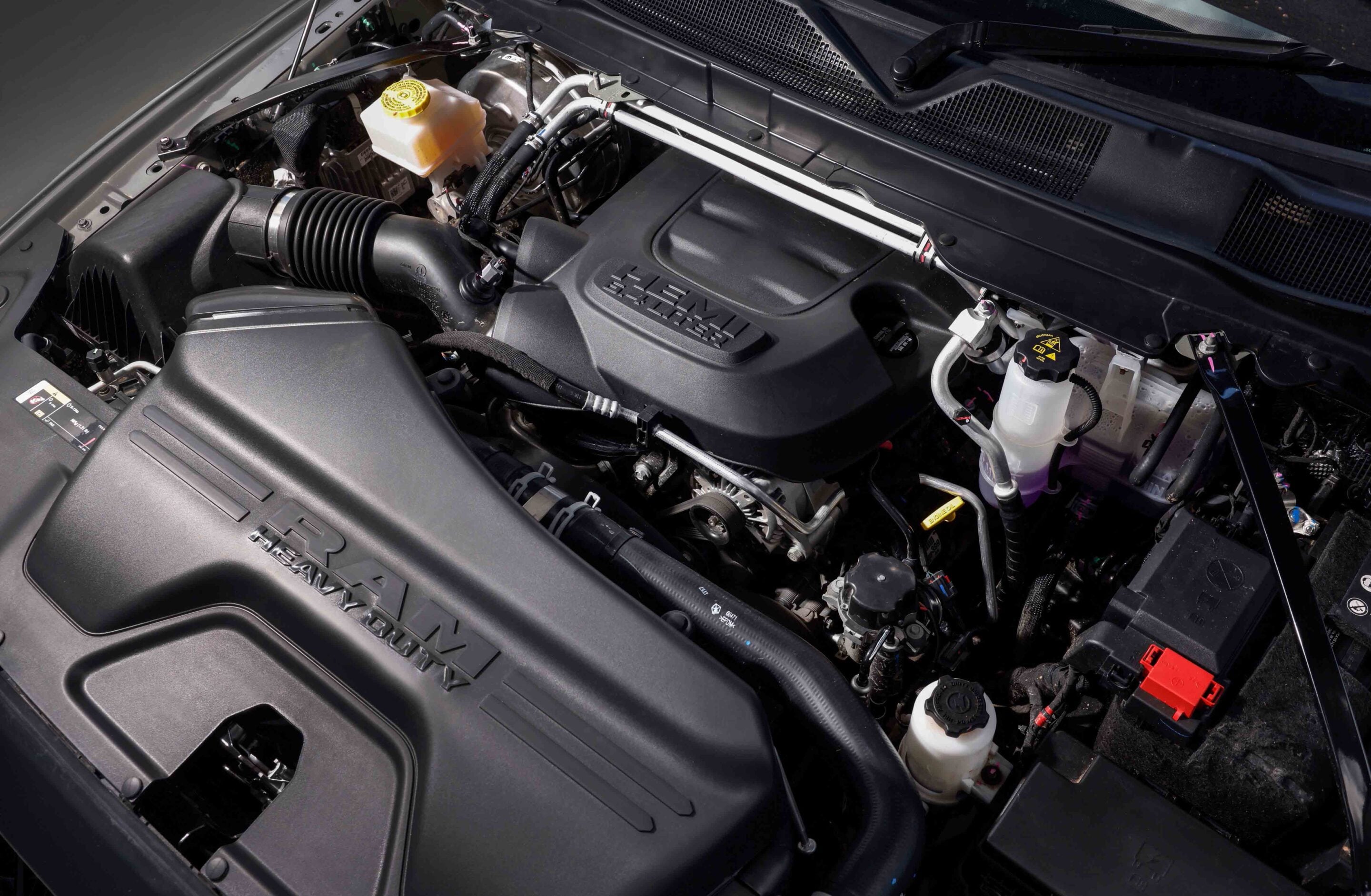 Under-hood view of a vehicle's engine bay with a black HEMI engine cover, coolant reservoir, hoses and metal pipes visible across the bay.