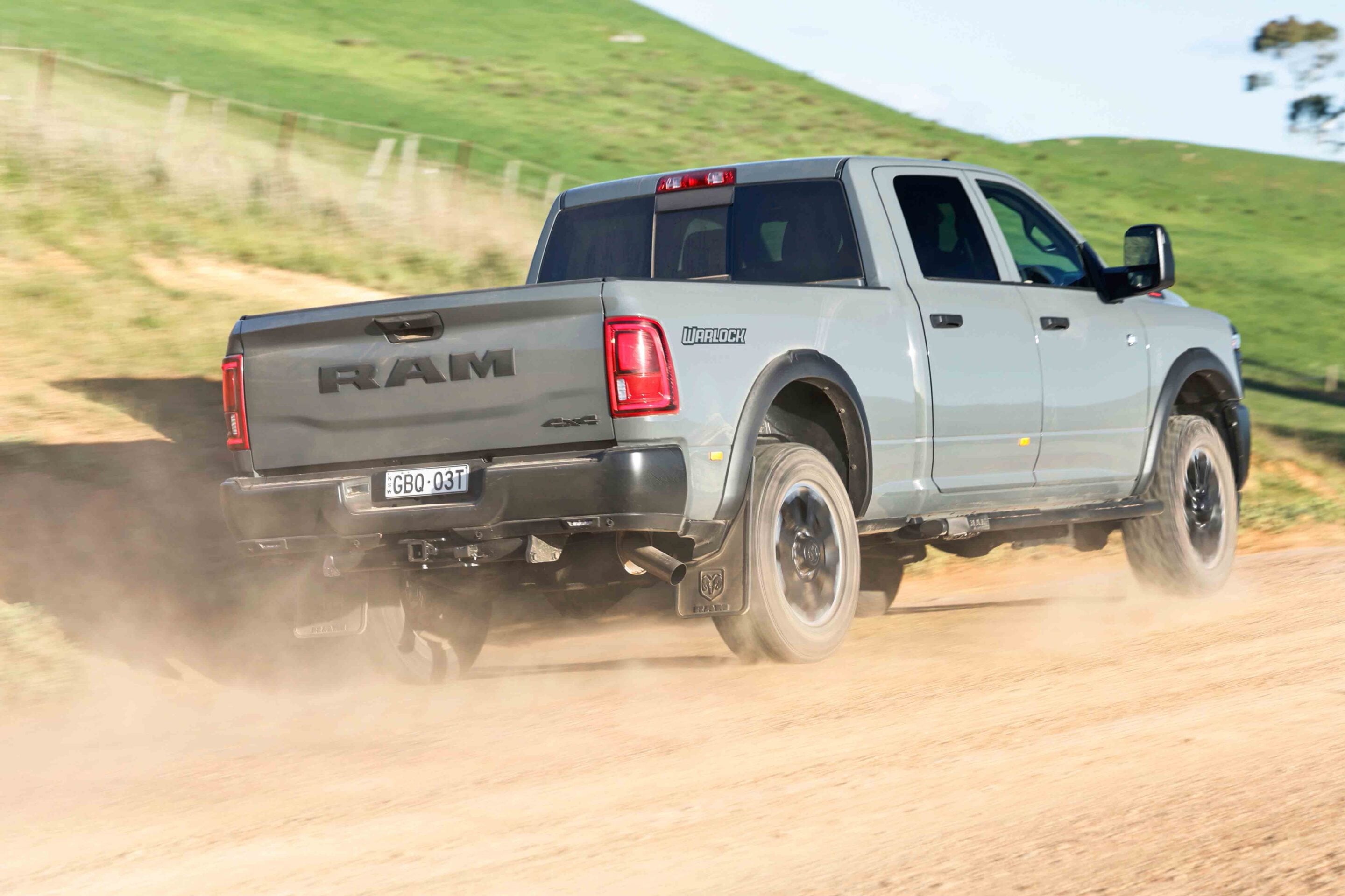 Rear three-quarter view of a gray RAM pickup speeding along a dusty rural road with green hills in the background and dirt clouds around the wheels.
