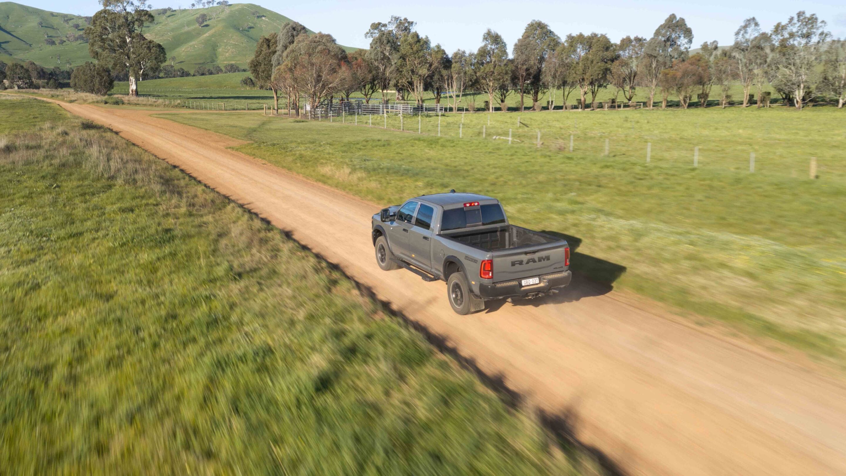Grey RAM pickup driving along a dirt country road with green fields and hills in the background.