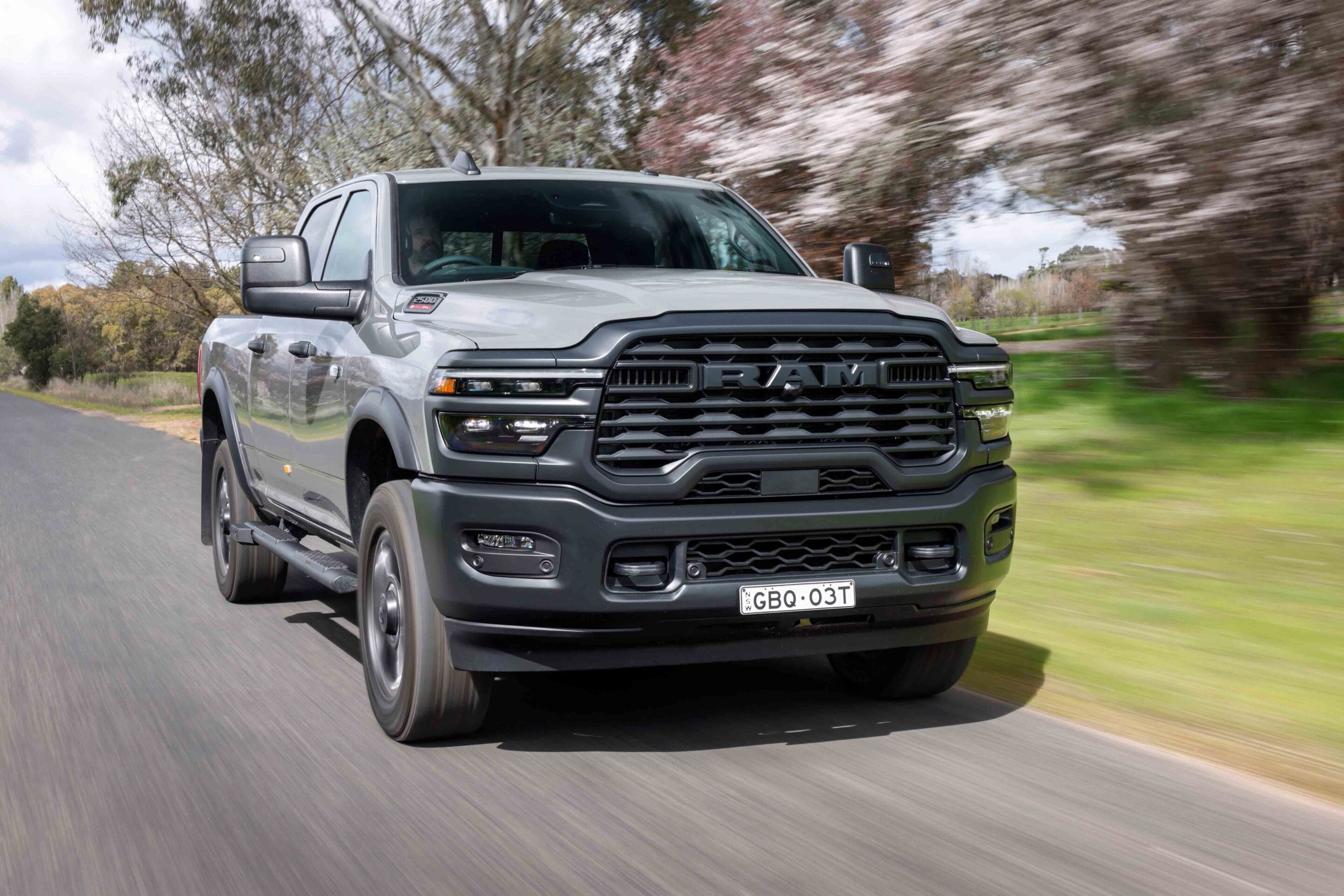 Front view of a silver RAM pickup driving toward the camera on a blurred country road, trees in the background.