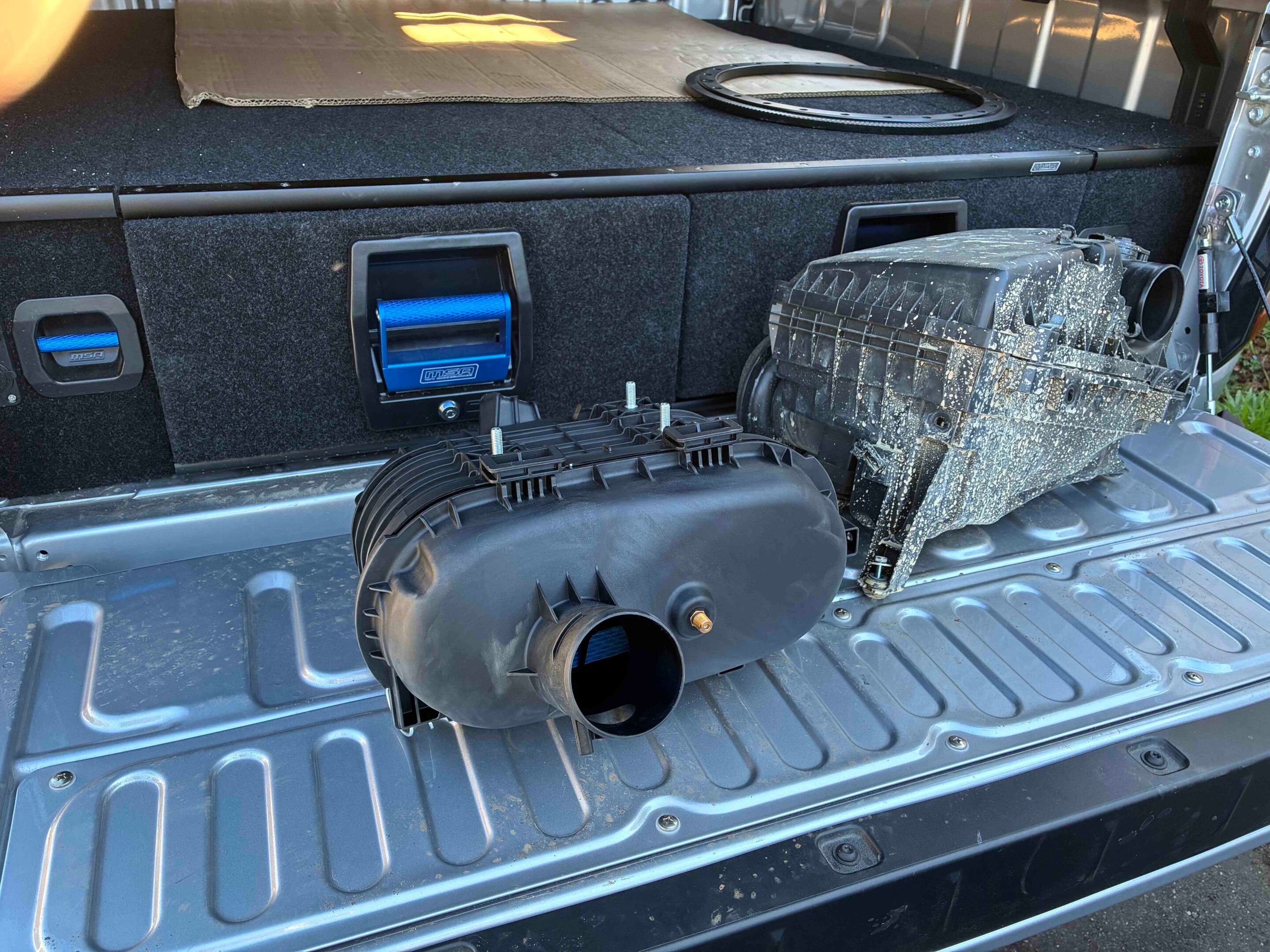 Open car trunk showing a large circular air intake duct and a blue rectangular component mounted in a recessed side compartment, with black carpeted panels and exposed hardware nearby.