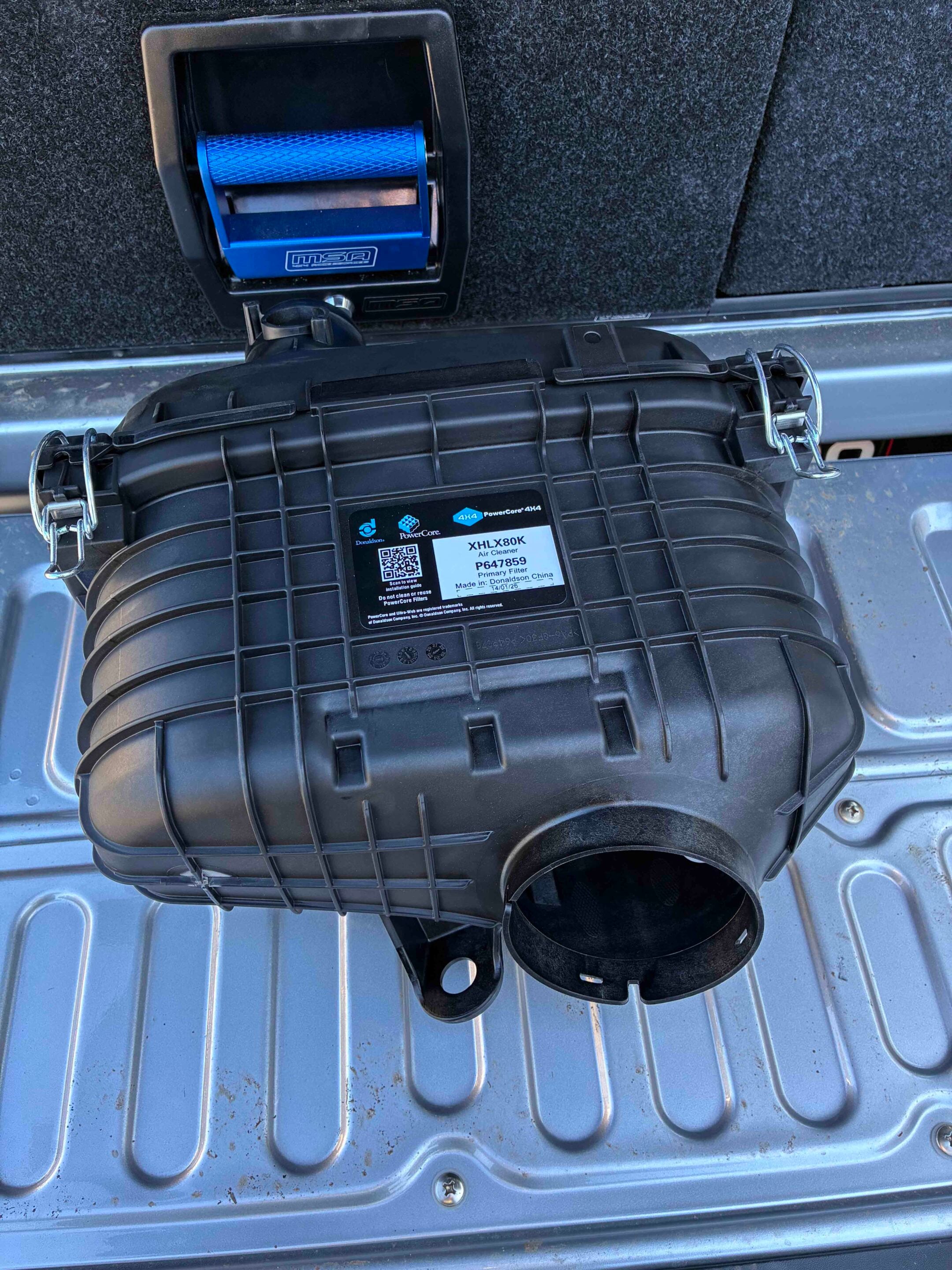 Blue rectangular air filter housing with a round duct, resting on a textured car trunk floor.