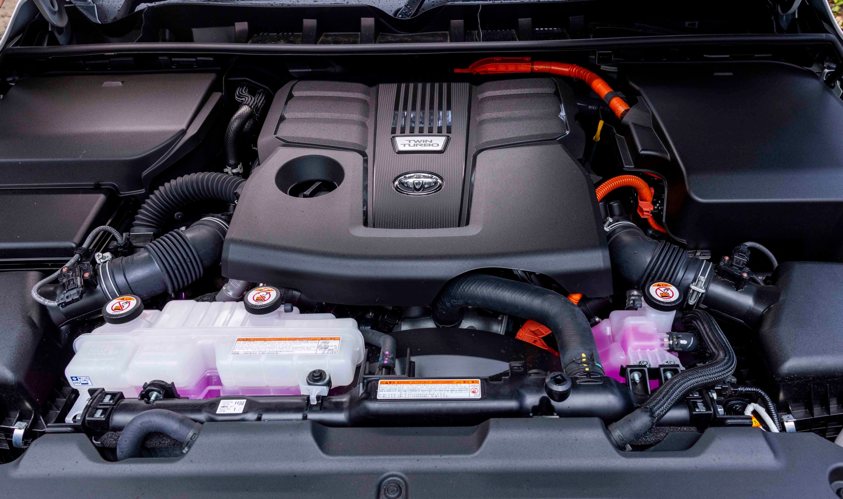 Car engine bay featuring a central Twin Turbo cover with Toyota emblem and surrounding hoses and battery packs.
