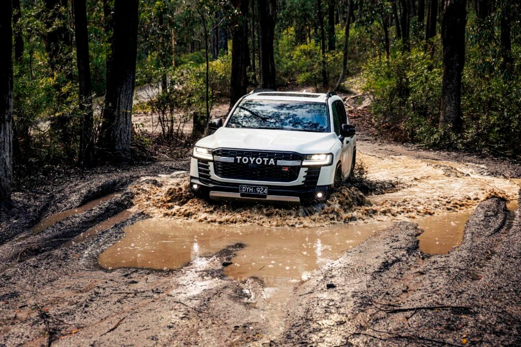 White Toyota SUV driving through a muddy forest trail with large puddles and splashes.