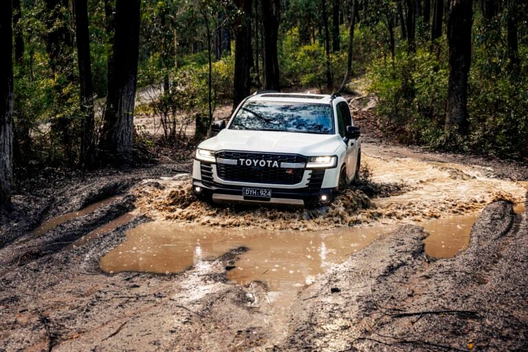 White Toyota SUV driving through a muddy forest trail with large puddles and splashes.