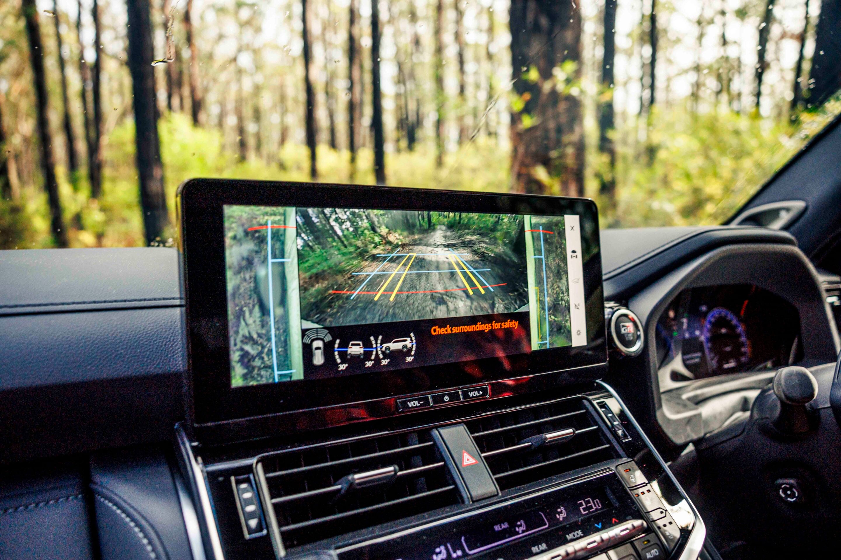 In-car center screen shows rear-camera view of a forested road with parking guidelines.