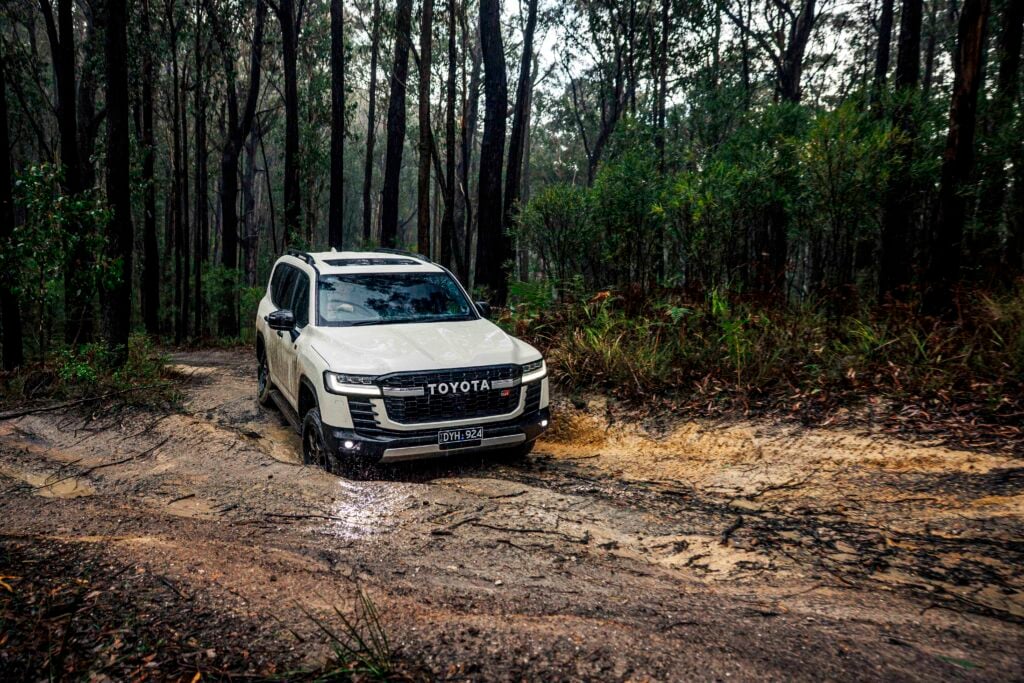 White Toyota SUV parked on a muddy forest trail with dense trees surrounding it, headlights on.
