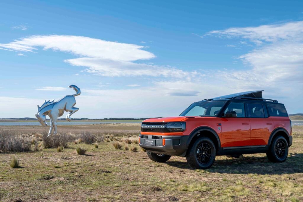 Orange Ford Bronco parked in a dry, grassy field with a metallic dinosaur sculpture nearby against a blue sky.