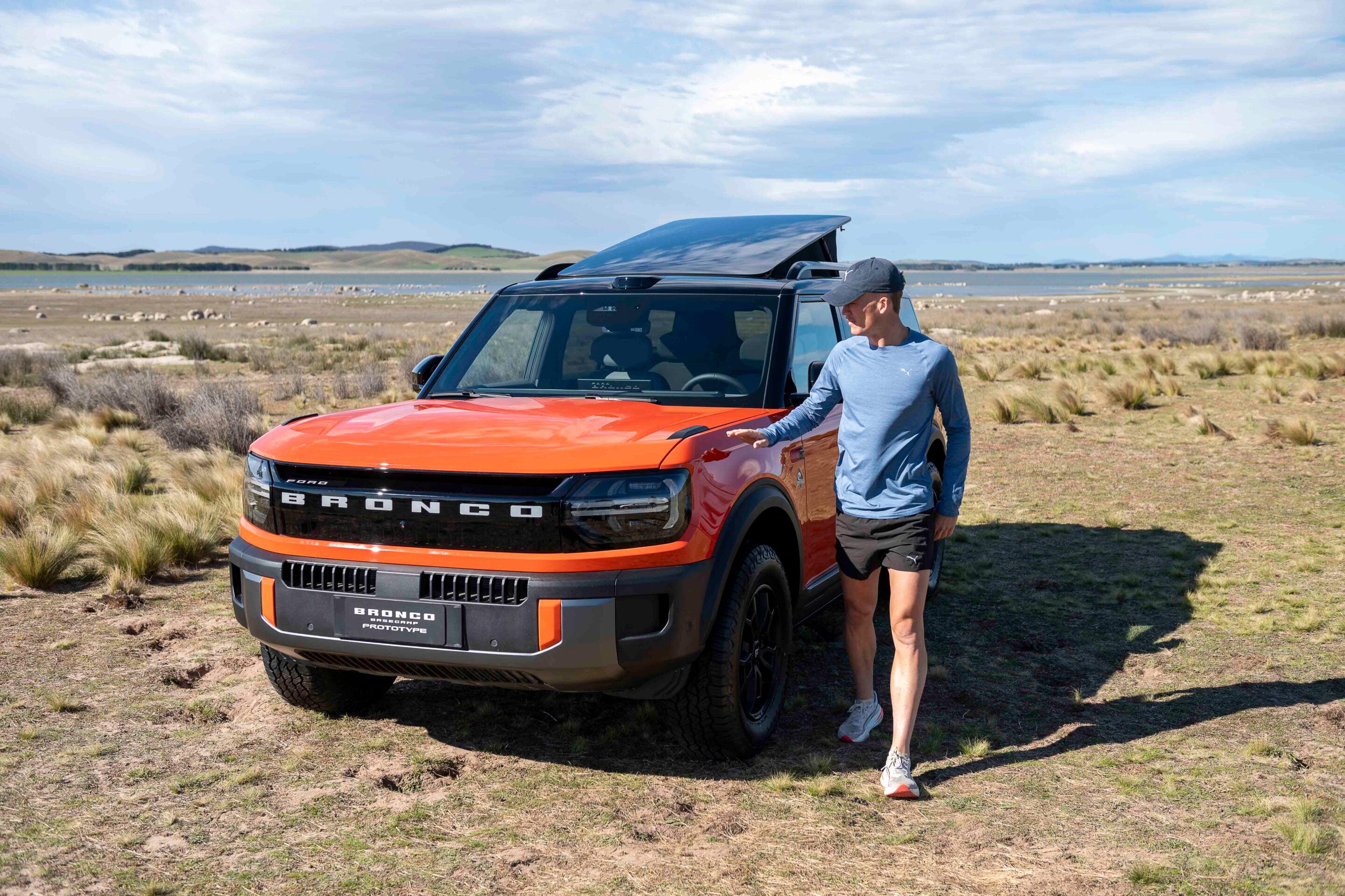 Man in a blue long-sleeve shirt and cap standing beside an orange Bronco SUV with the sunroof open in a dry, grassy landscape near a lake.