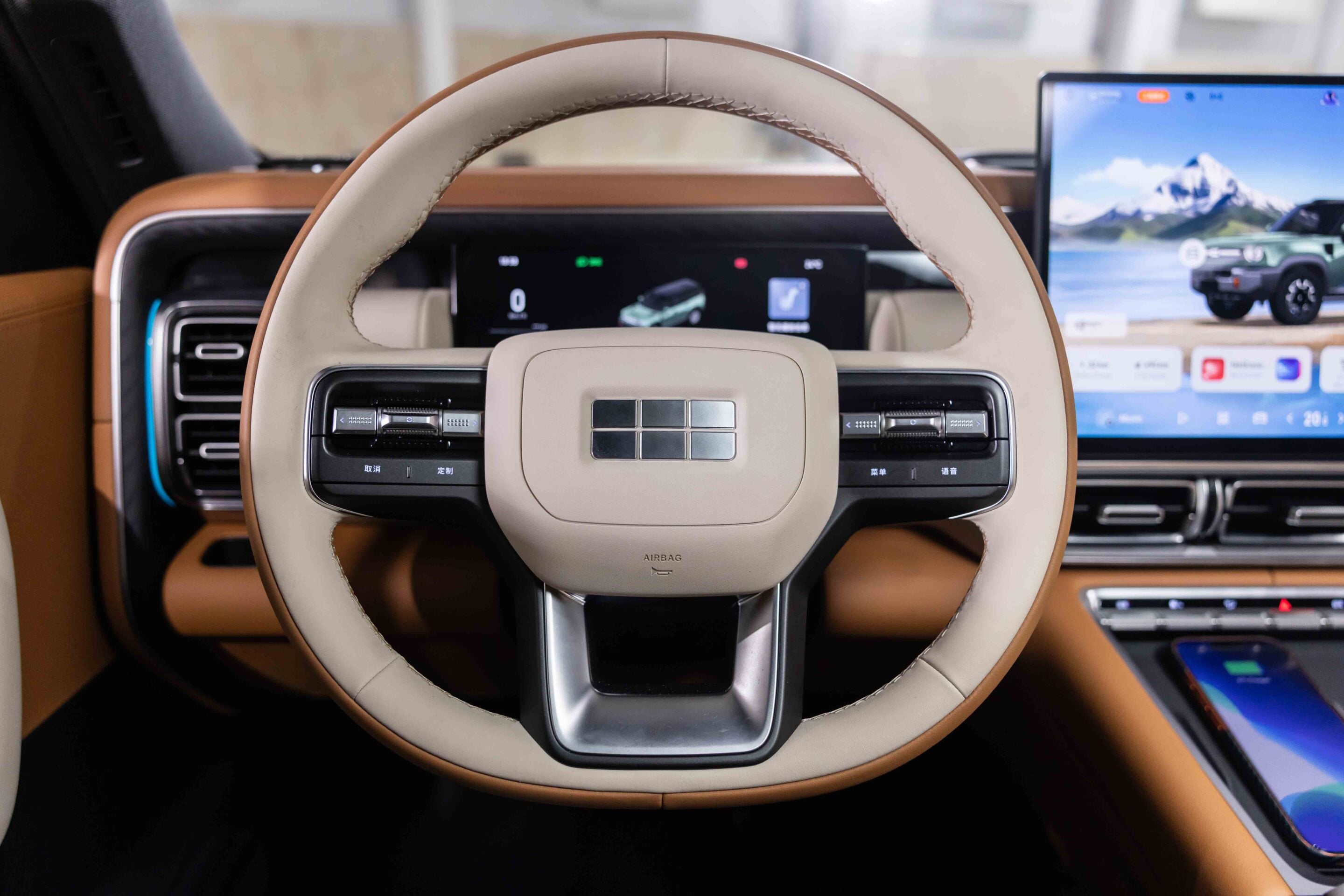 Beige leather steering wheel with central airbag and digital dashboard, plus a large infotainment screen showing a car and mountain scenery in the background.