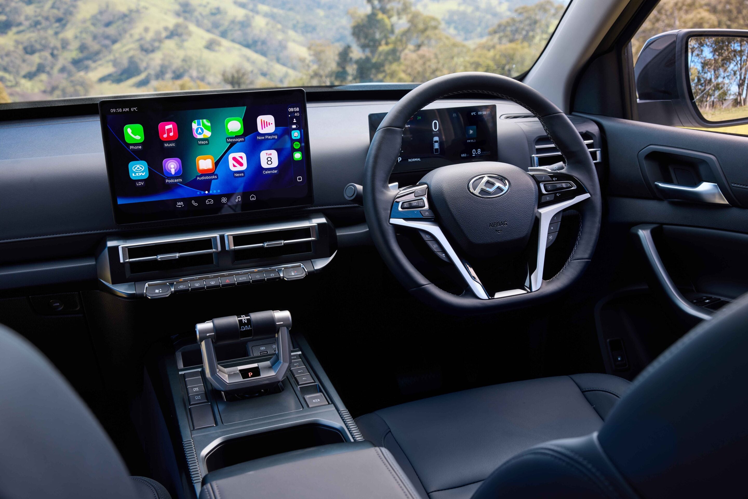 Interior of a car with a large touchscreen infotainment display on the dashboard next to the steering wheel and a digital instrument cluster behind it, plus exterior hills visible through the windshield.