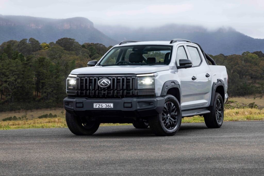 Front three-quarter view of a white pickup truck parked on a road, with a forest and mountains in the background.