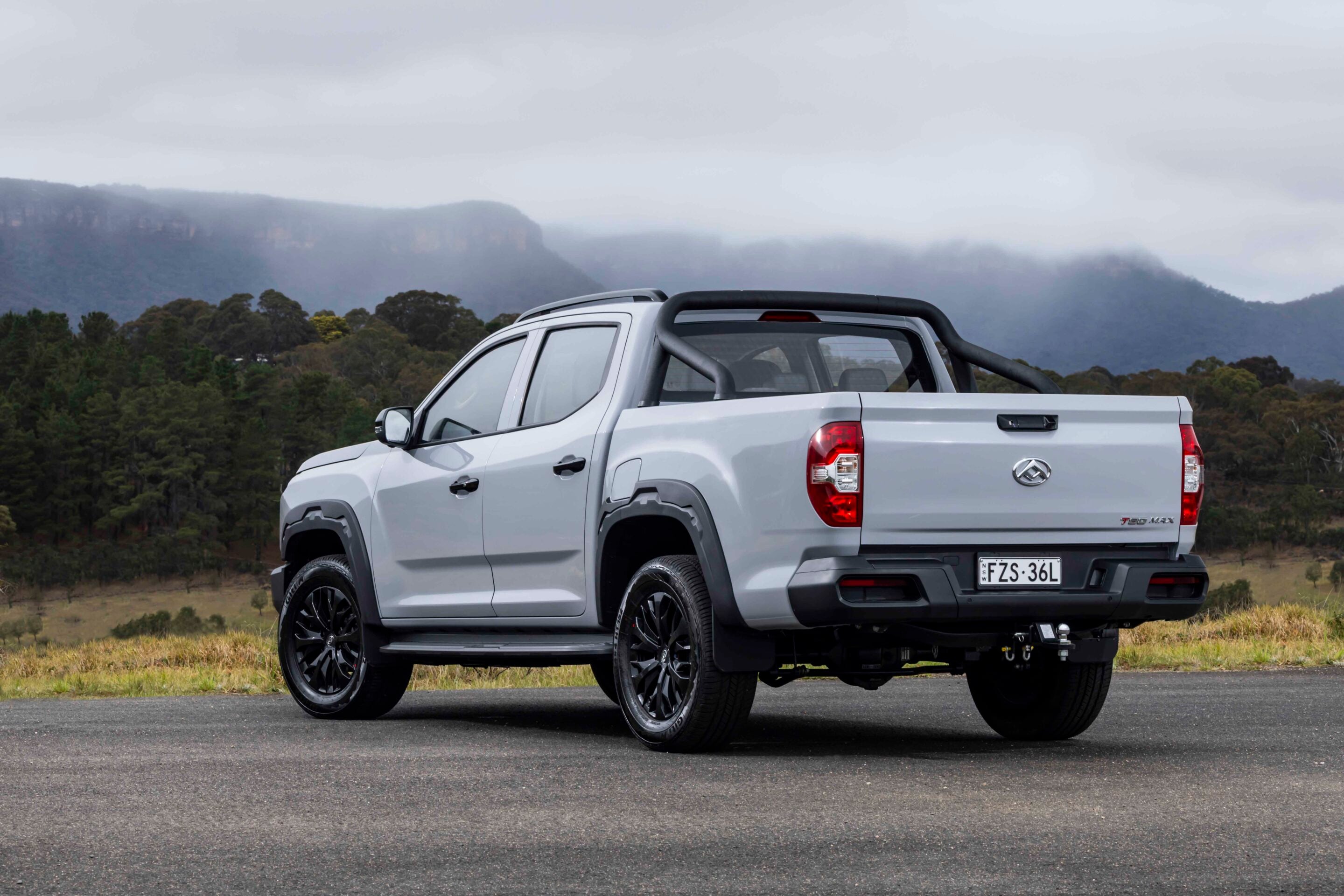 Rear three-quarter view of a silver pickup truck with a roll bar, parked on a paved surface against a backdrop of misty mountains and a forested landscape.