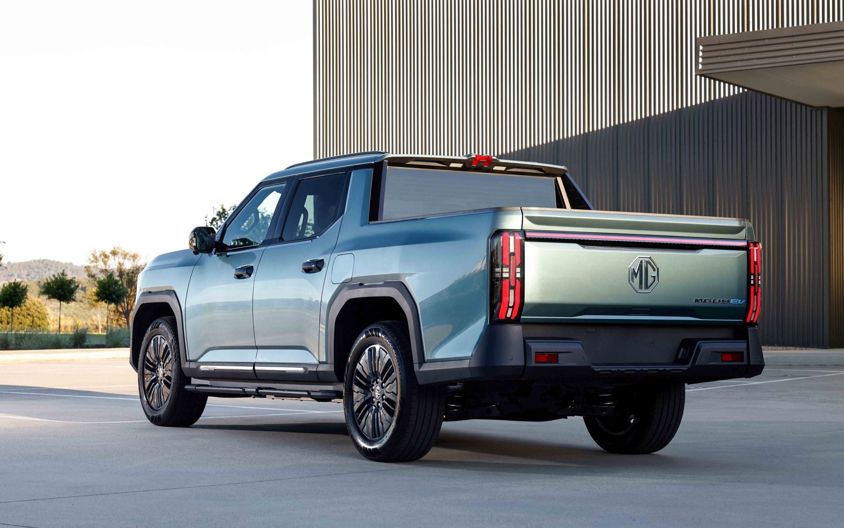 Rear three-quarter view of a metallic blue MG electric pickup parked outdoors near a corrugated metal building, showing the MG badge on the tailgate and red taillights.