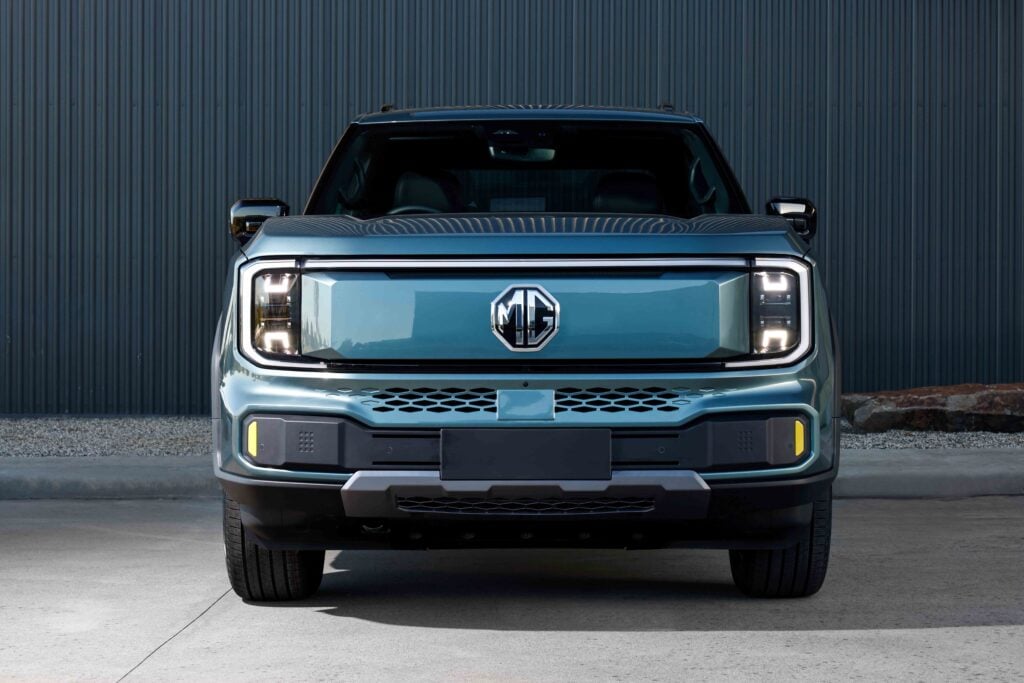 Front view of a blue MG pickup truck facing the camera against a corrugated metal wall.