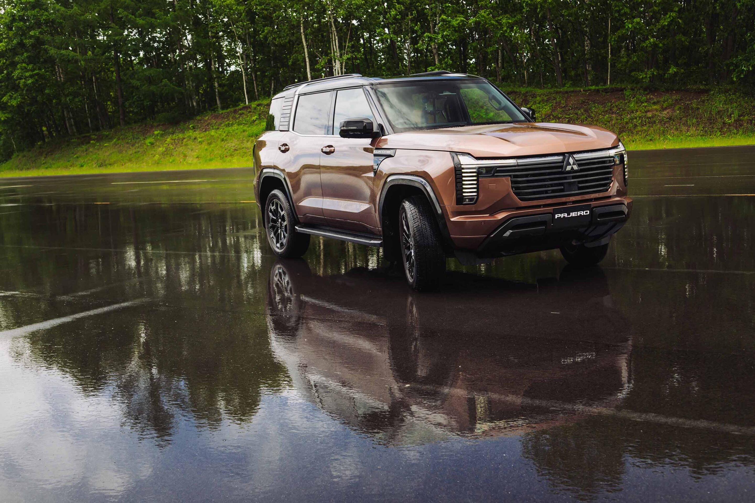 Brown Mitsubishi Pajero SUV parked on a wet road, reflections in puddles and a forest backdrop.