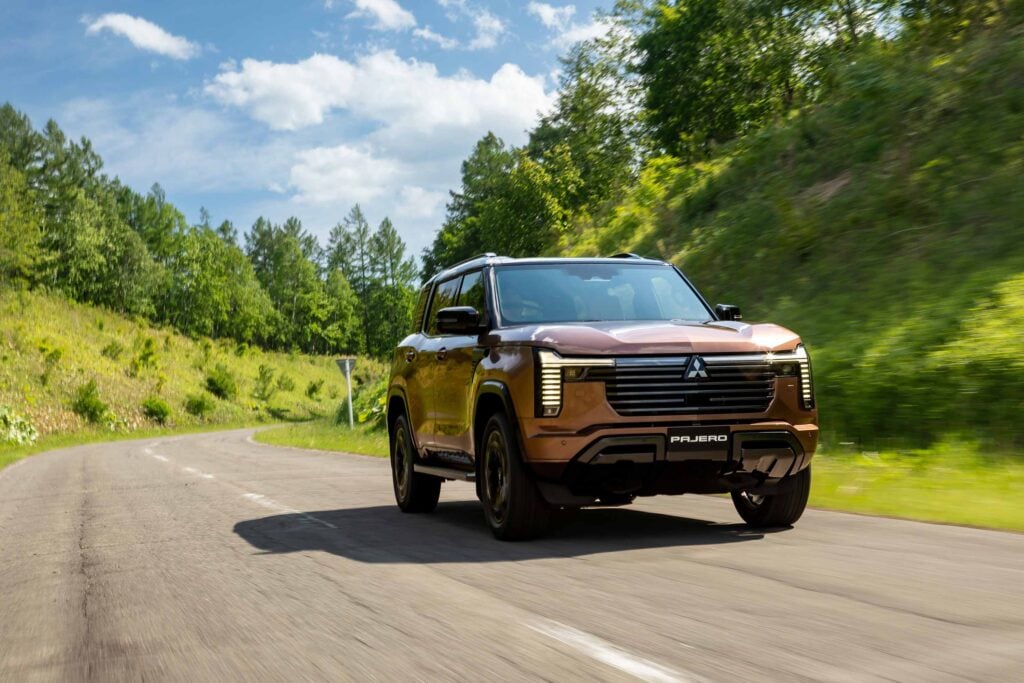 Brown Mitsubishi Pajero driving on a winding country road with trees and blue sky above.