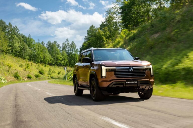 Brown Mitsubishi Pajero driving on a winding country road with trees and blue sky above.