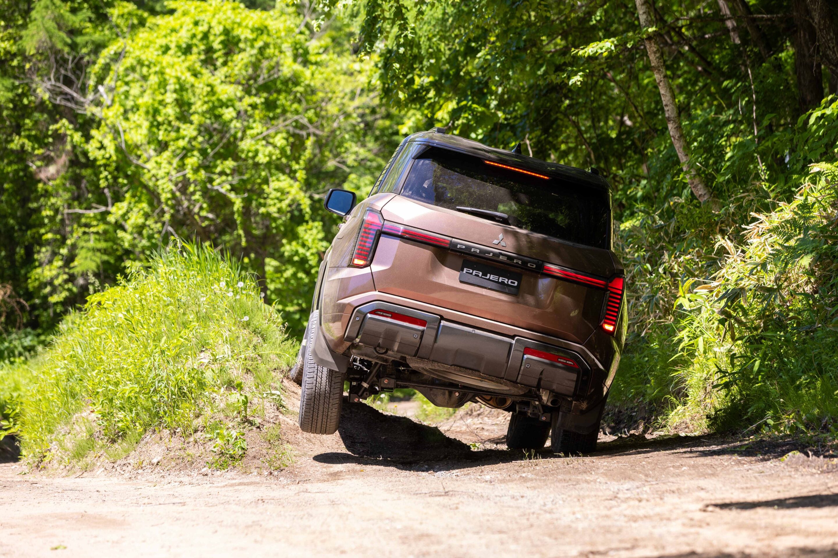 Brown Mitsubishi Pajero driving off-road on a dirt path surrounded by dense green forest, with the rear of the SUV visible and wheels angled over uneven terrain.