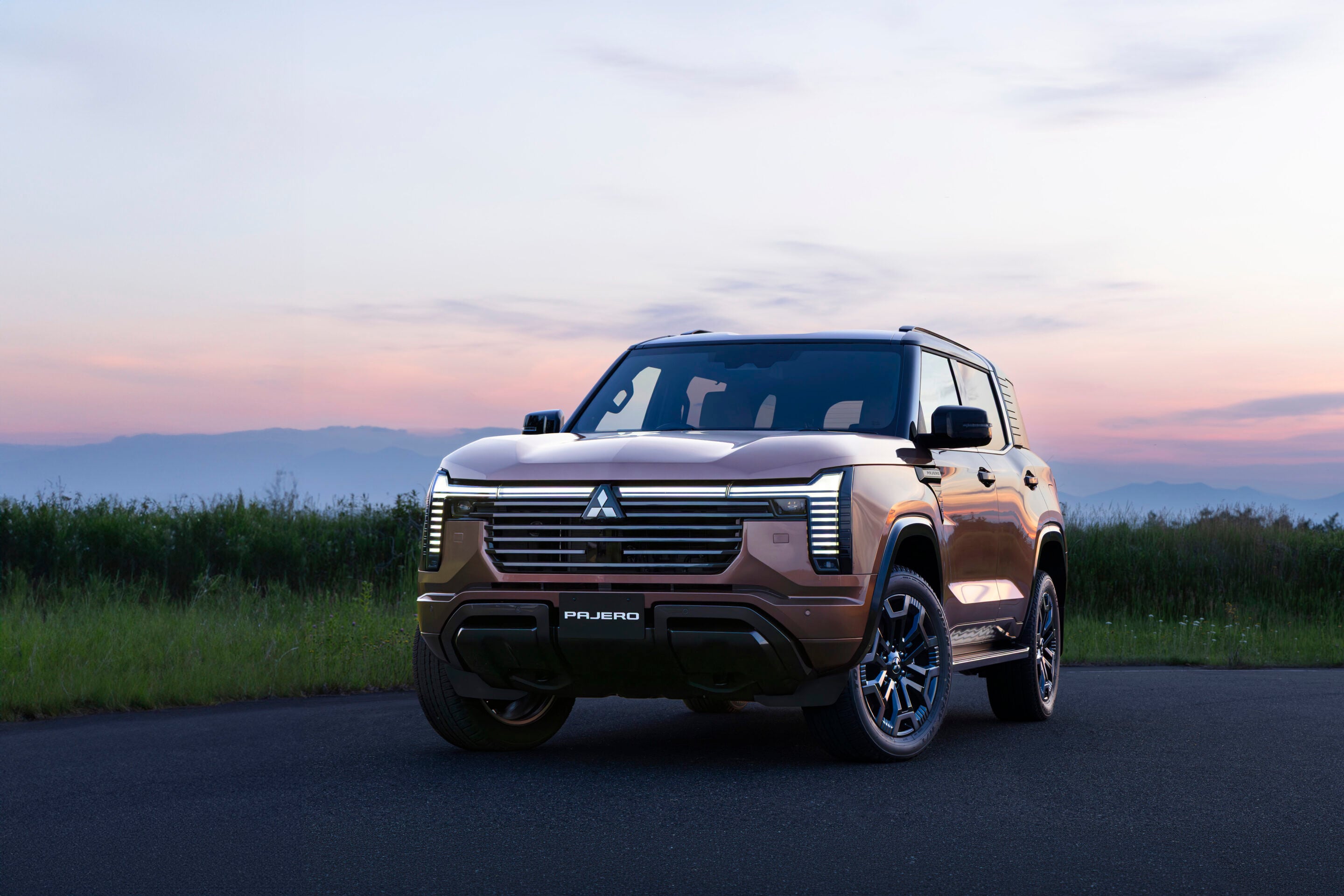 Front-right view of a bronze Mitsubishi Pajero SUV parked on a paved road at sunset, with a grassy field and distant hills in the background.