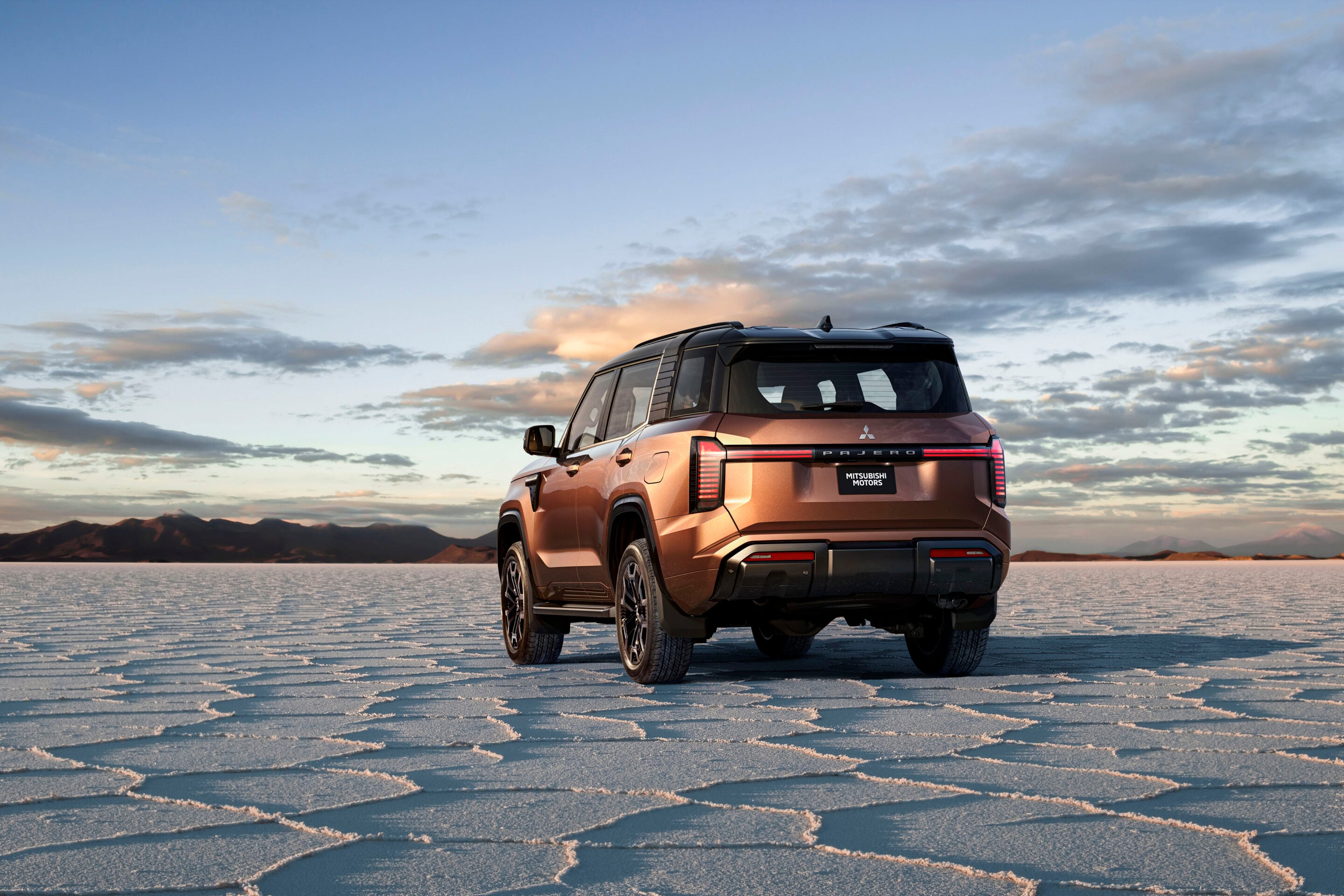 Orange Mitsubishi Pajero SUV seen from the rear on a cracked salt flat with mountains in the distance at sunset.