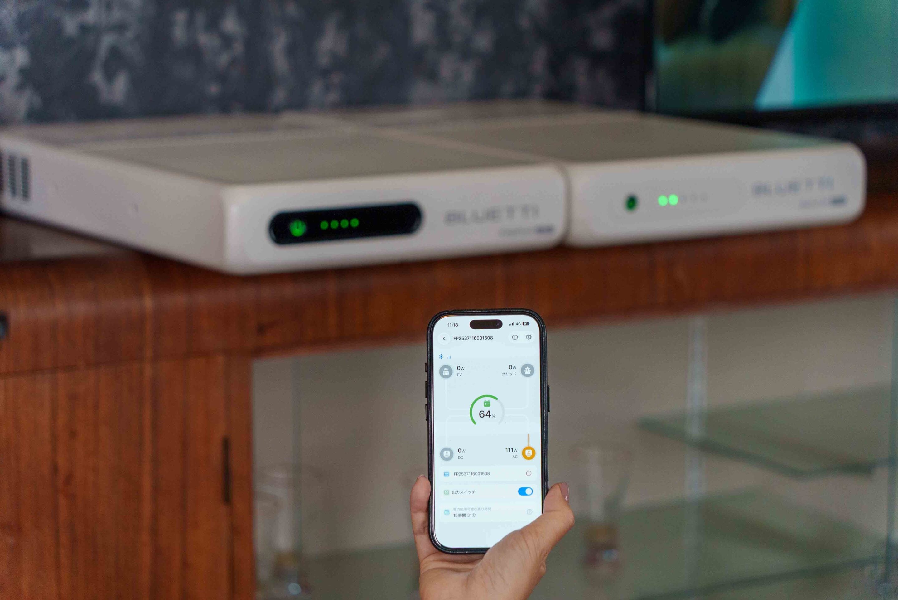 A person holds a smartphone displaying a dashboard with a circular 64% gauge, with two white network devices on a wooden shelf in the background.