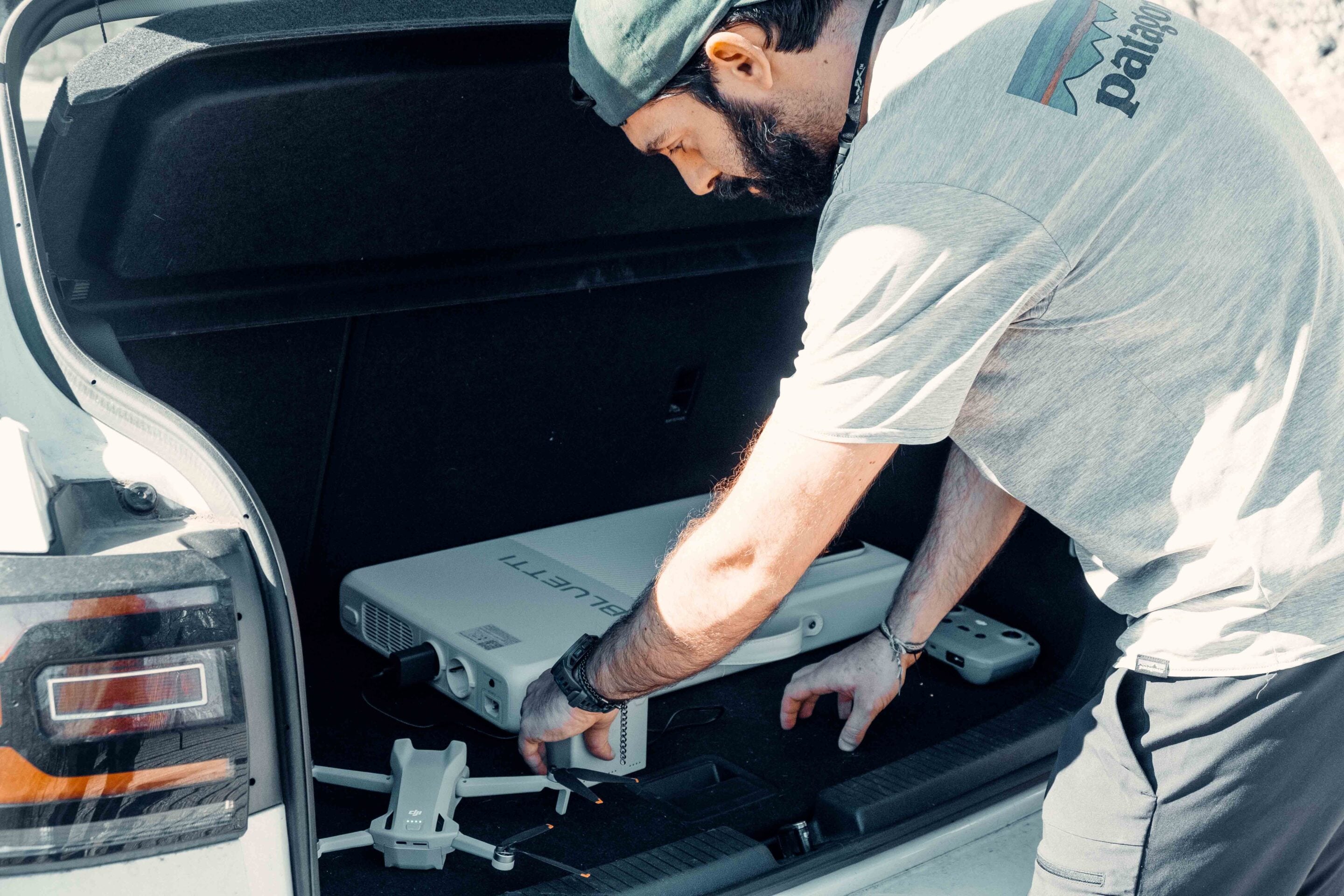 Man in a gray shirt and cap bending into an open car trunk, handling a large drone and its controller inside the vehicle's cargo area.