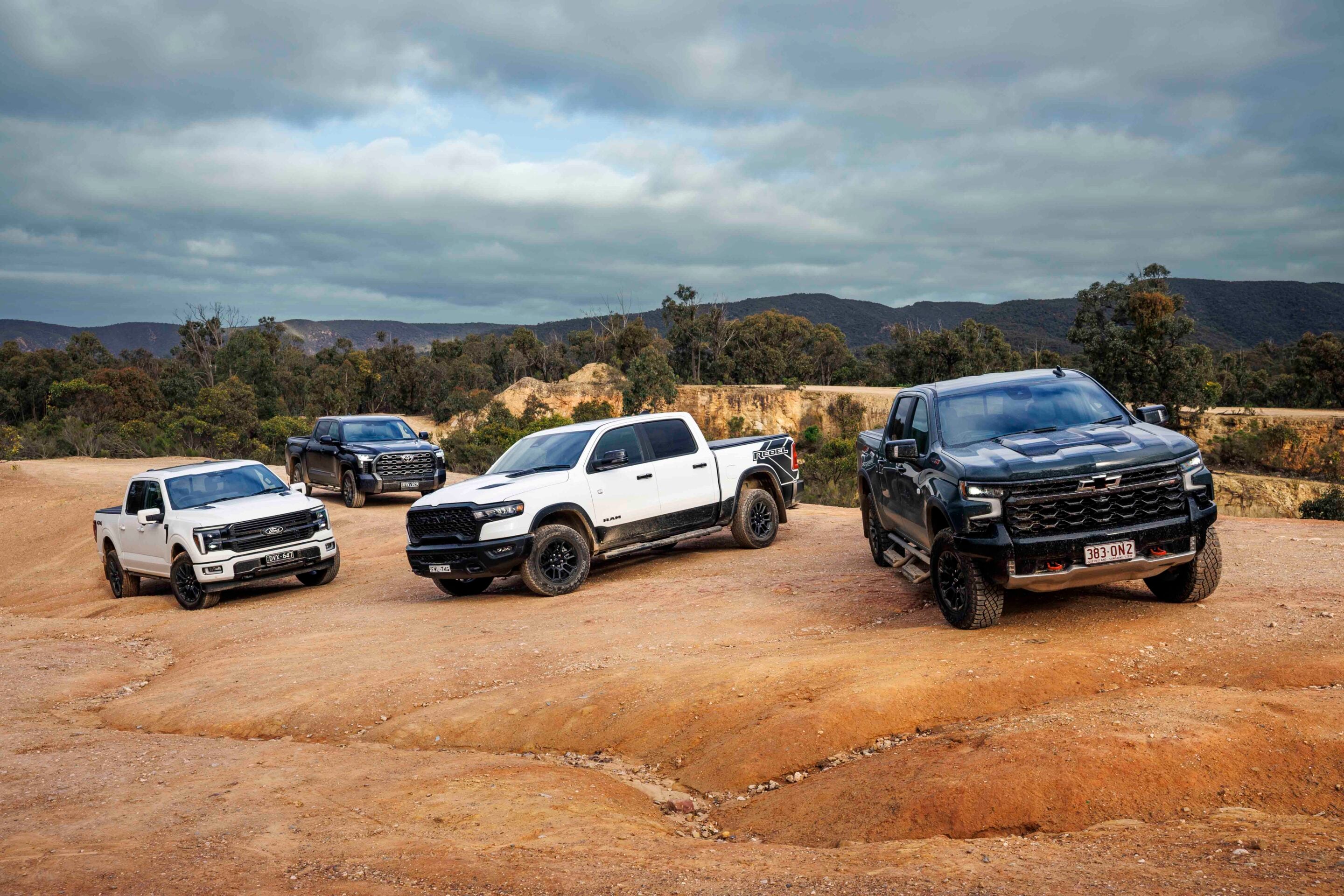 Group of pickup trucks parked on a dusty dirt lot with hills and cloudy sky in the background