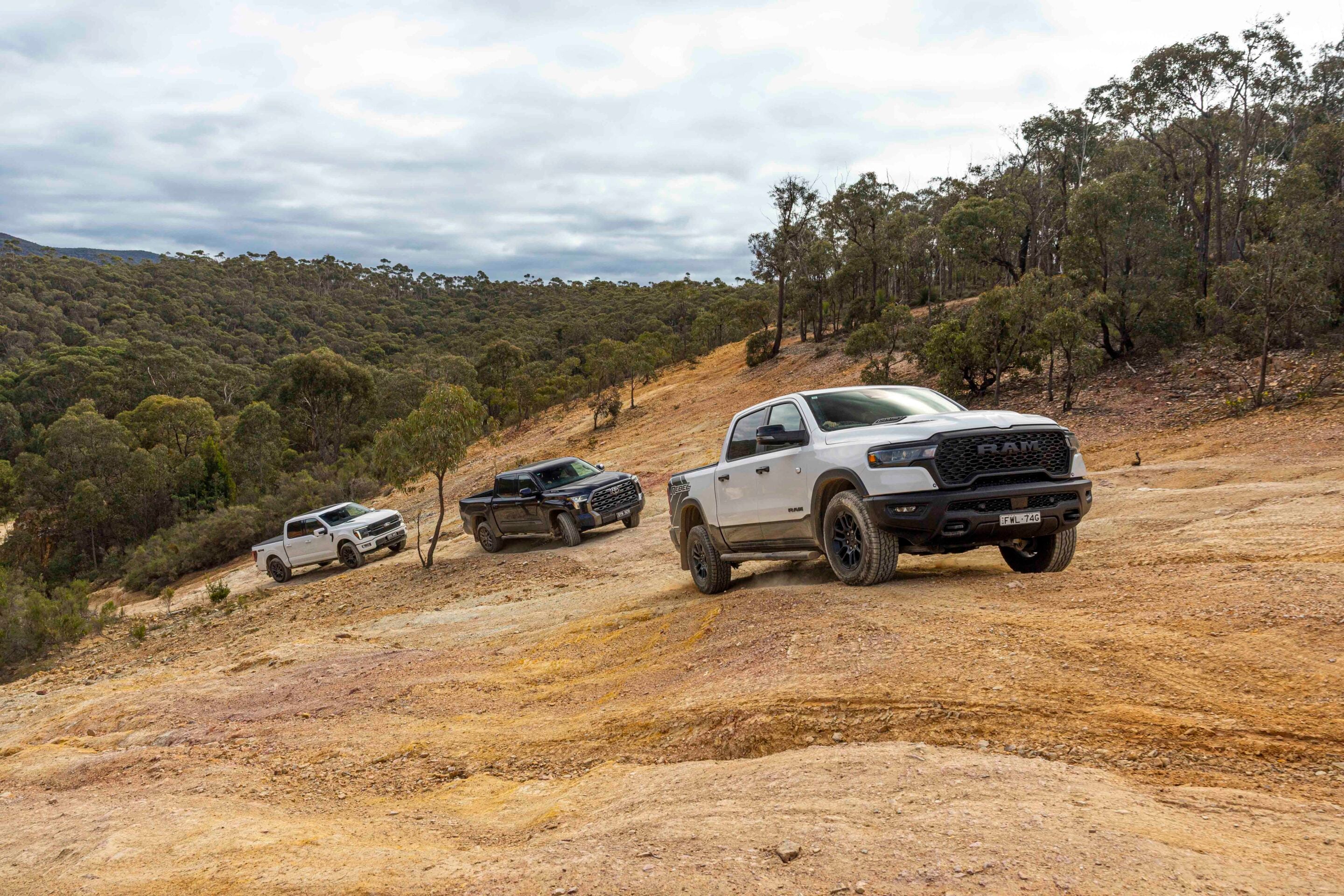 Off-road pickup trucks parked on a dusty hillside with a scrubby forest and cloudy sky in the background.