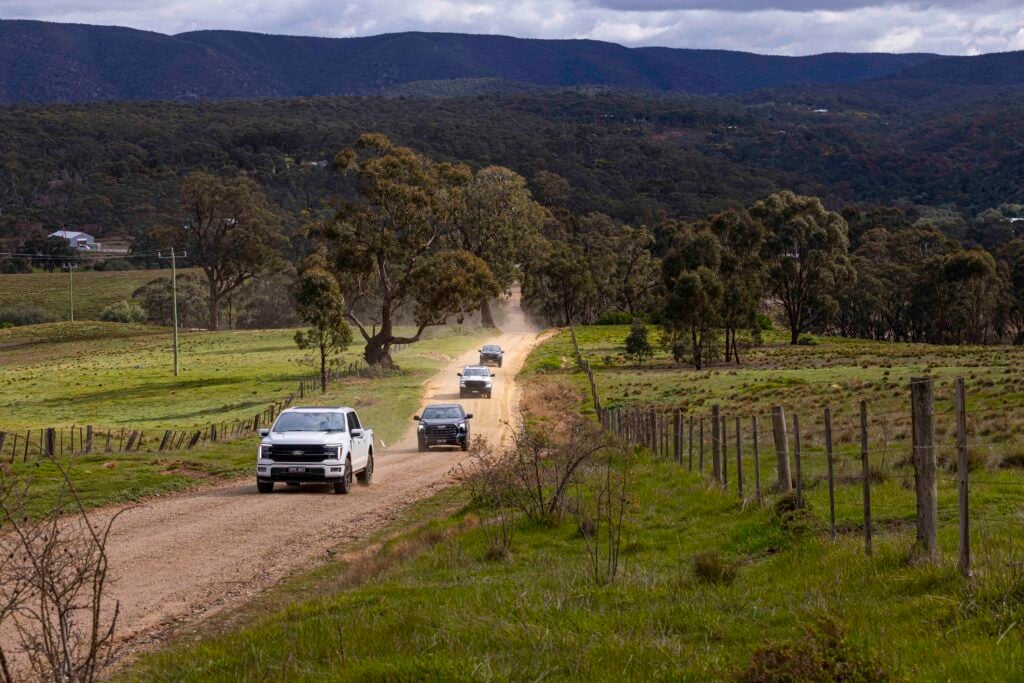 Dusty rural dirt road with a line of cars driving, fenced fields and trees, and distant hills and mountains.