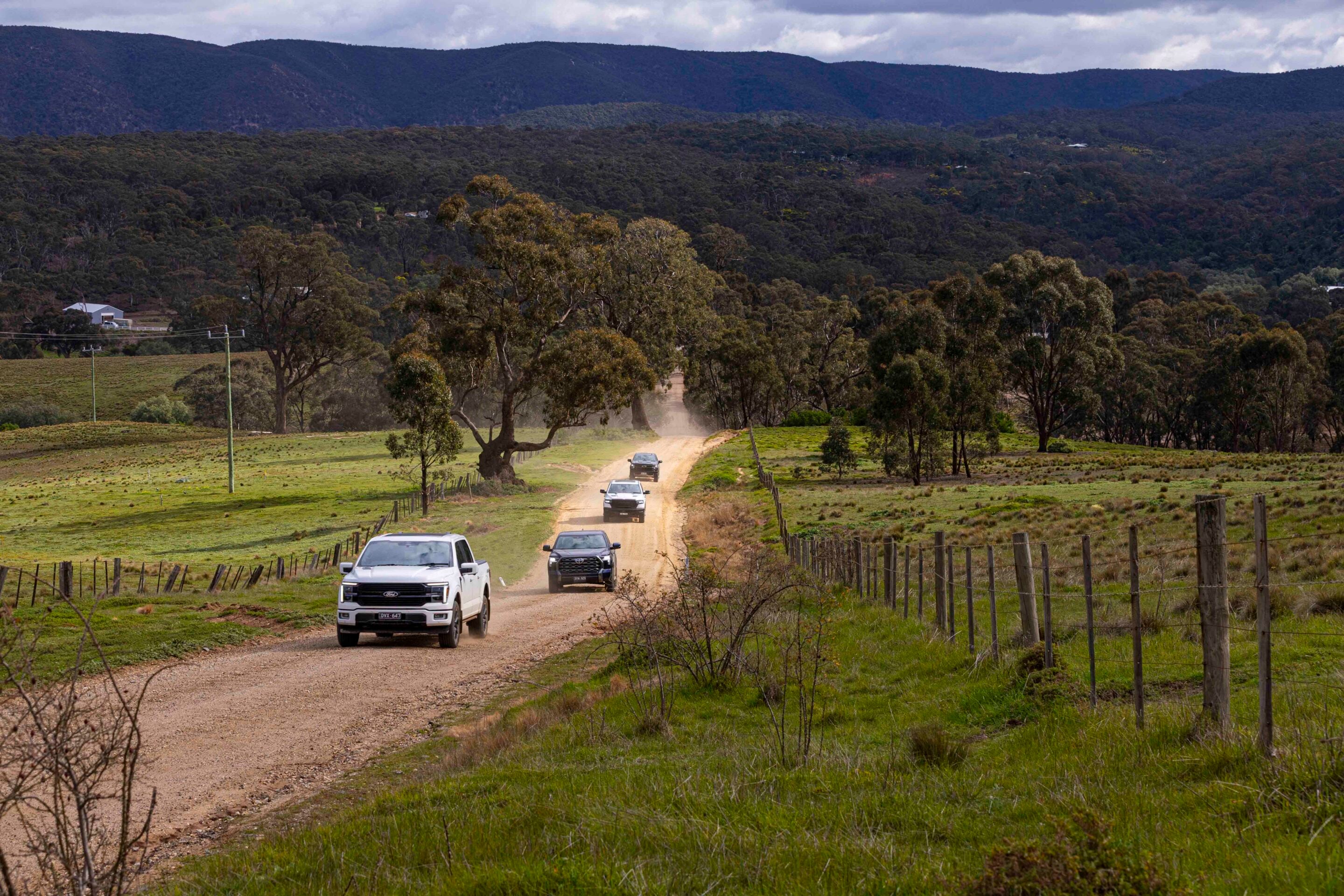 Dusty rural dirt road with a line of cars driving, fenced fields and trees, and distant hills and mountains.