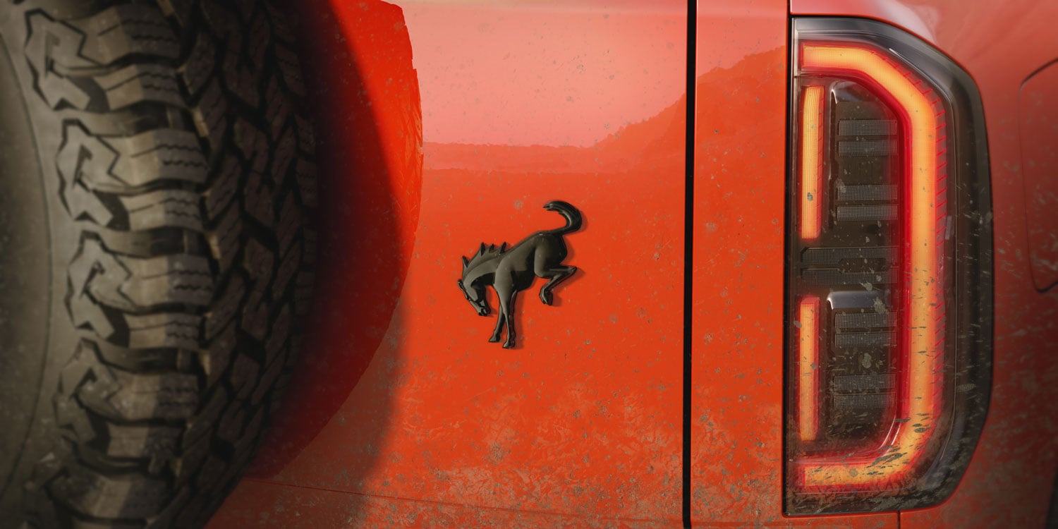 Orange vehicle side with a black Mustang emblem, a rugged left tire, and a red taillight on the right, muddy surface across the panel.