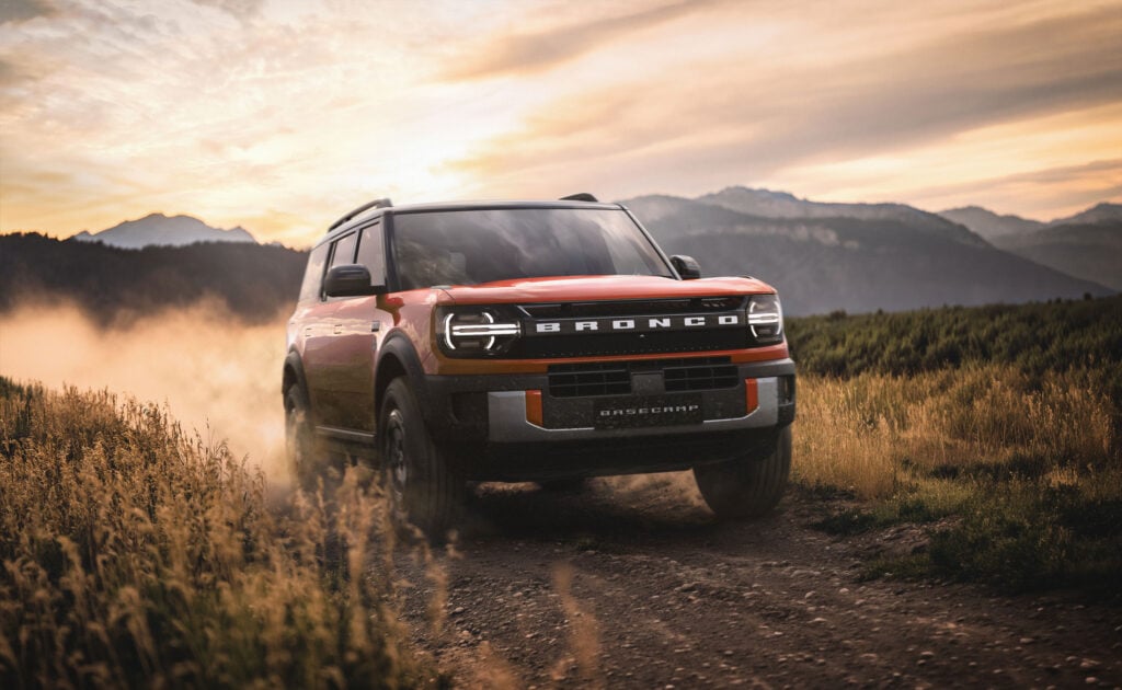 Red Ford Bronco SUV driving through a dusty dirt trail at sunset, with tall grasses and mountains in the background