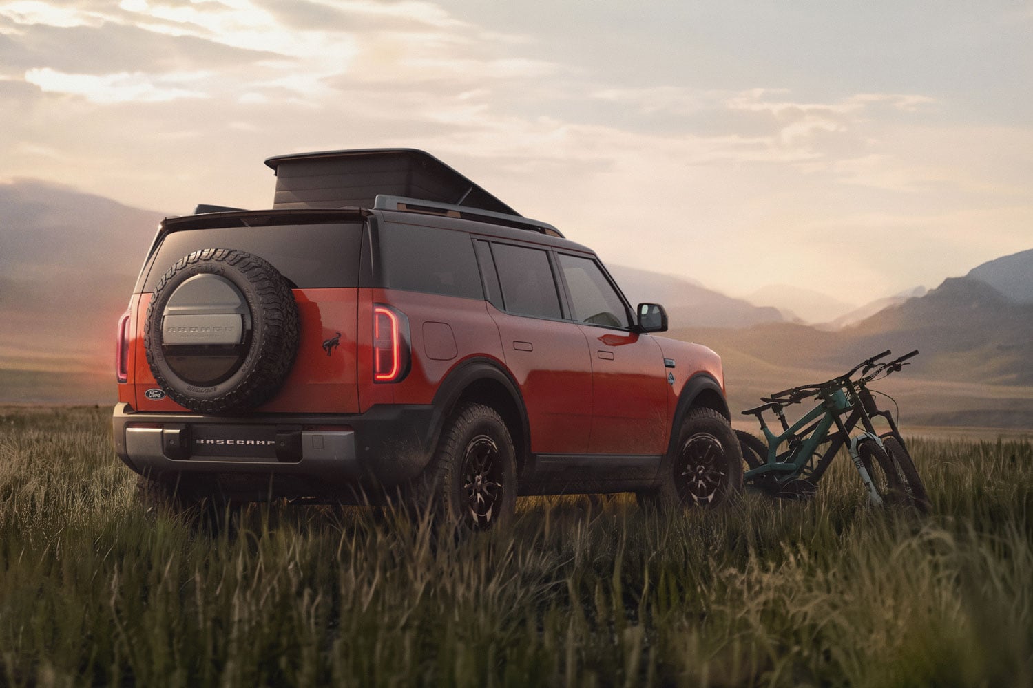Red Ford Bronco with a rear spare tire, parked in tall grass at sunset, with a rooftop tent and two bicycles resting beside it in a wide open landscape.
