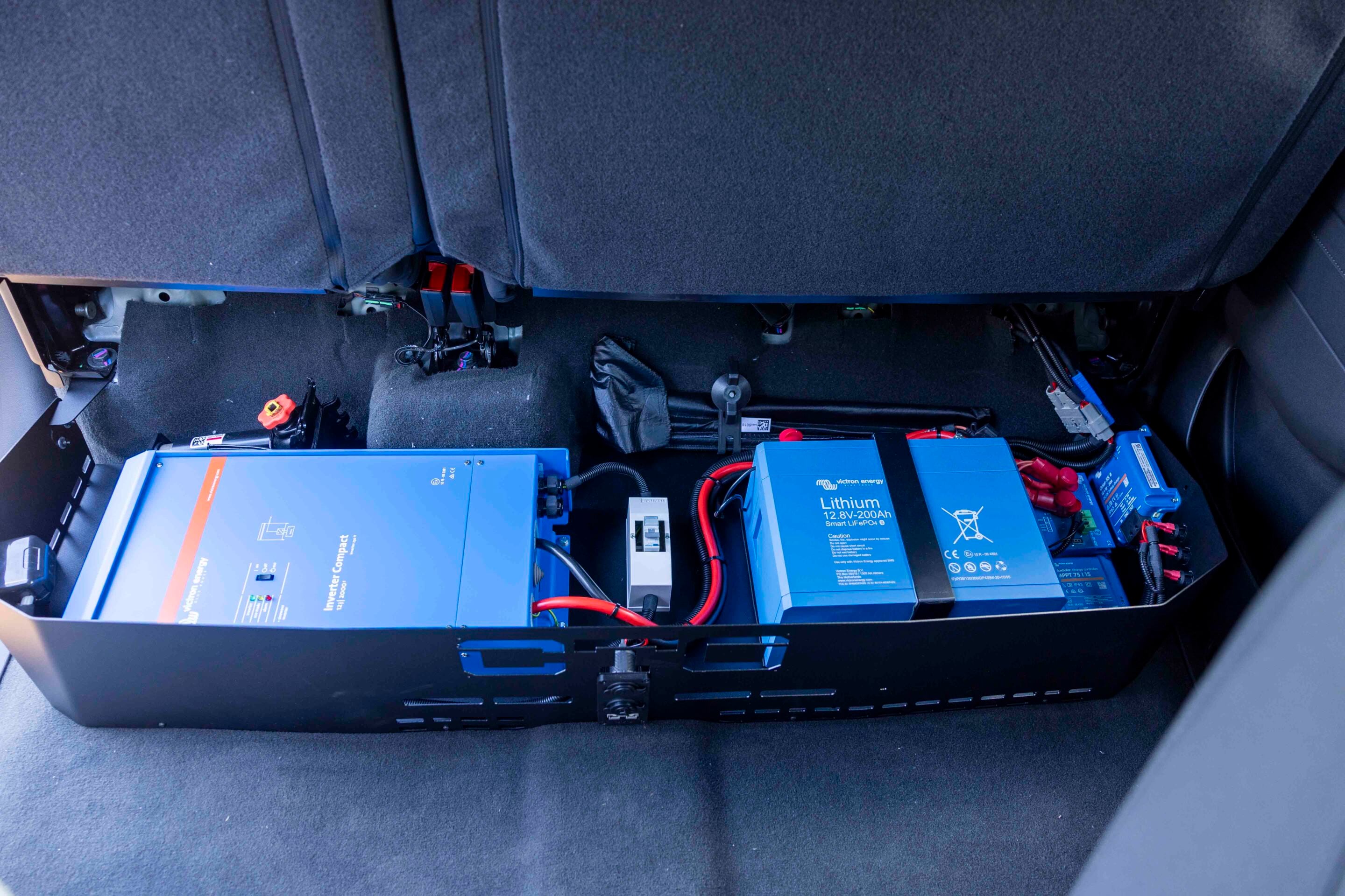 Blue lithium battery modules installed in a vehicle's rear cargo area, with red and black cables and a carpeted floor visible.
