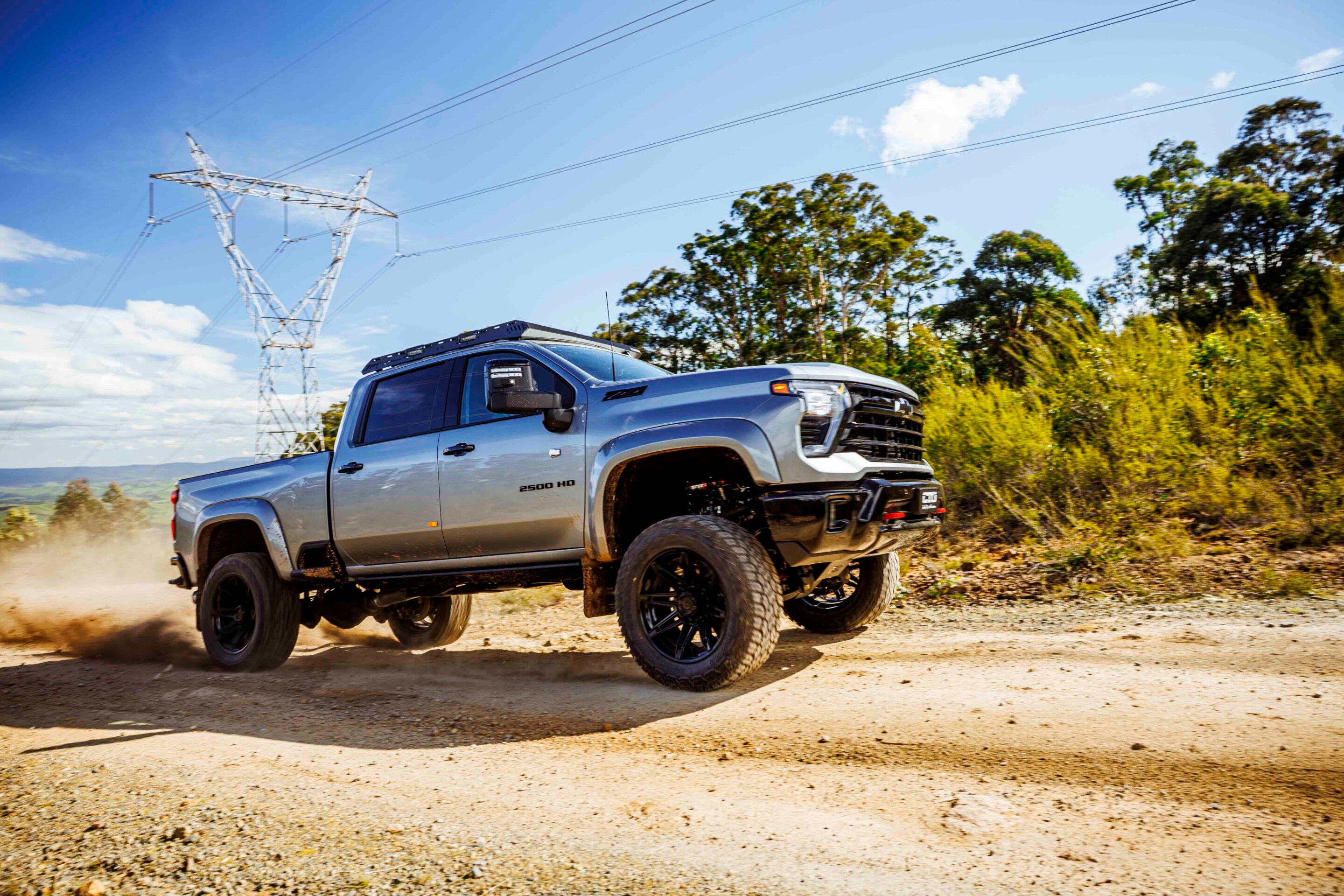 Silver lifted pickup truck kicking up dust on a dirt road with power lines and trees in the background.