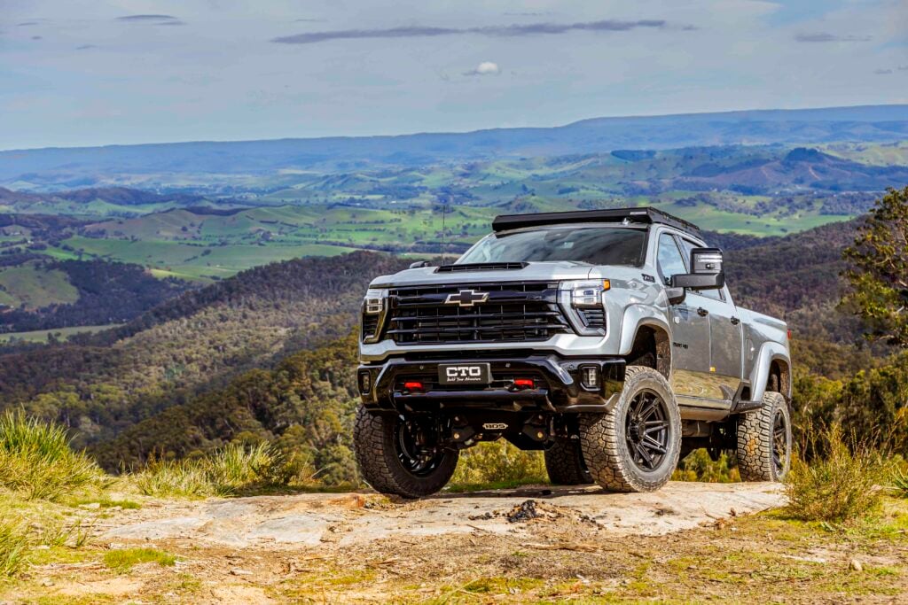 Silver Chevrolet pickup parked on a dirt trail with green rolling hills and blue sky in the background.